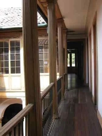 Indoor view of a hallway with wooden flooring, railing, and large windows on the left side, leading to a darker room at the end.