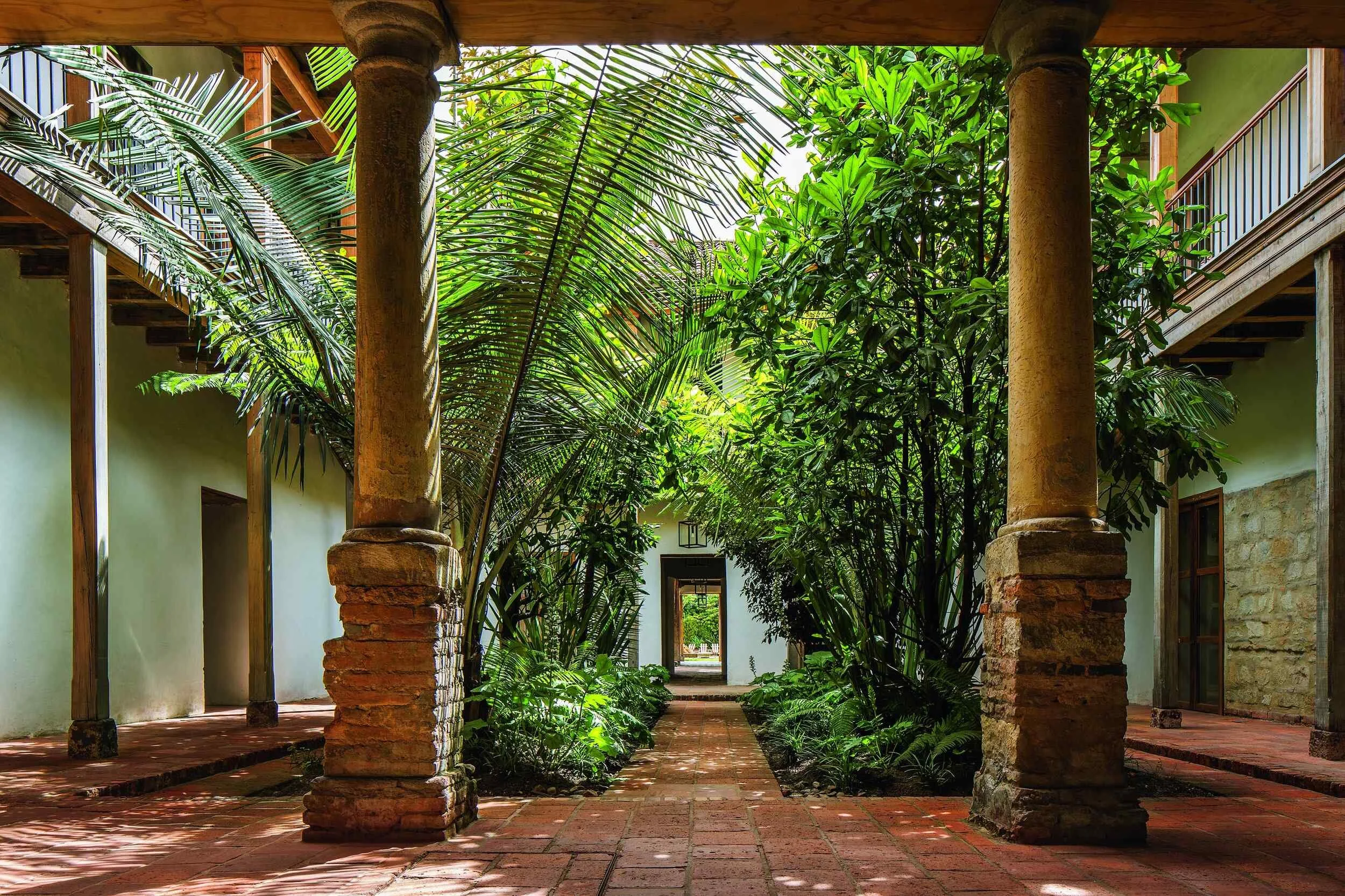 A lush green courtyard with brick paths, stone and wooden columns, enclosed by buildings with balconies and open doorways, shaded by large leafy plants.