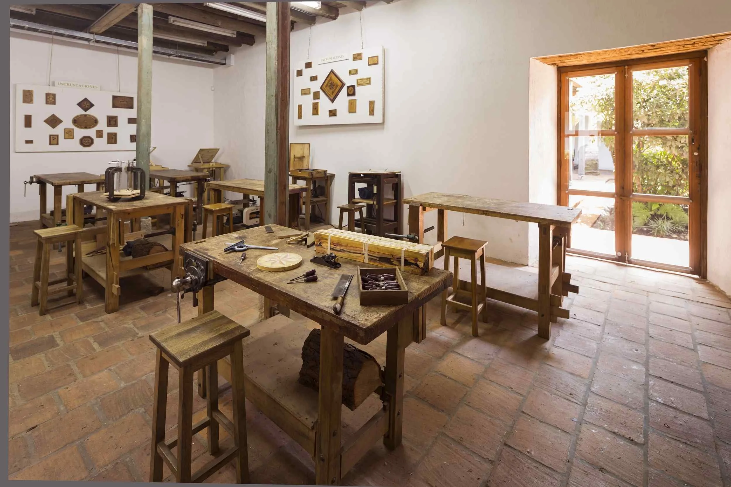 Workshop with wooden workbenches, stools, woodworking tools, and decorative plaques on the wall, illuminated by sunlight through a window.