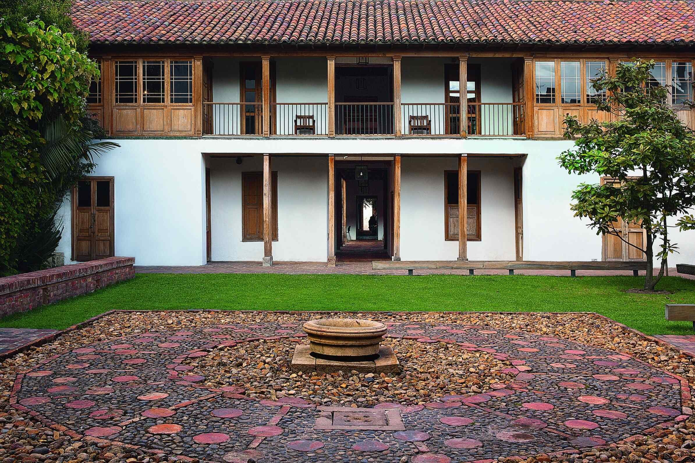 Front view of a two-story house with white walls, wooden accents, and a red-tiled roof, featuring a garden with a circular stone pathway and small fountain in the center.