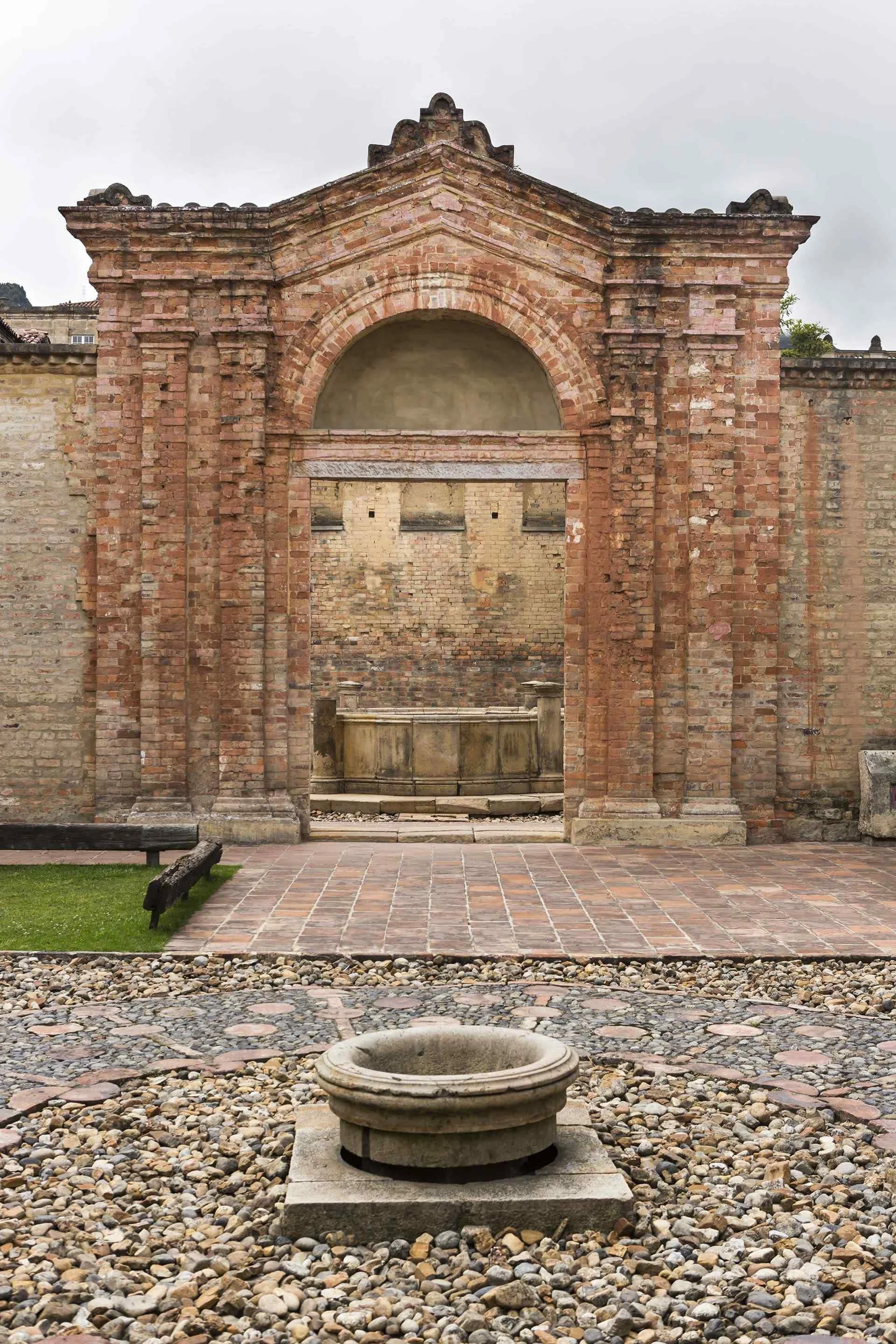 Ancient stone fountain in front of a historic brick wall with arched window and columns, on a paved courtyard with small rocks.