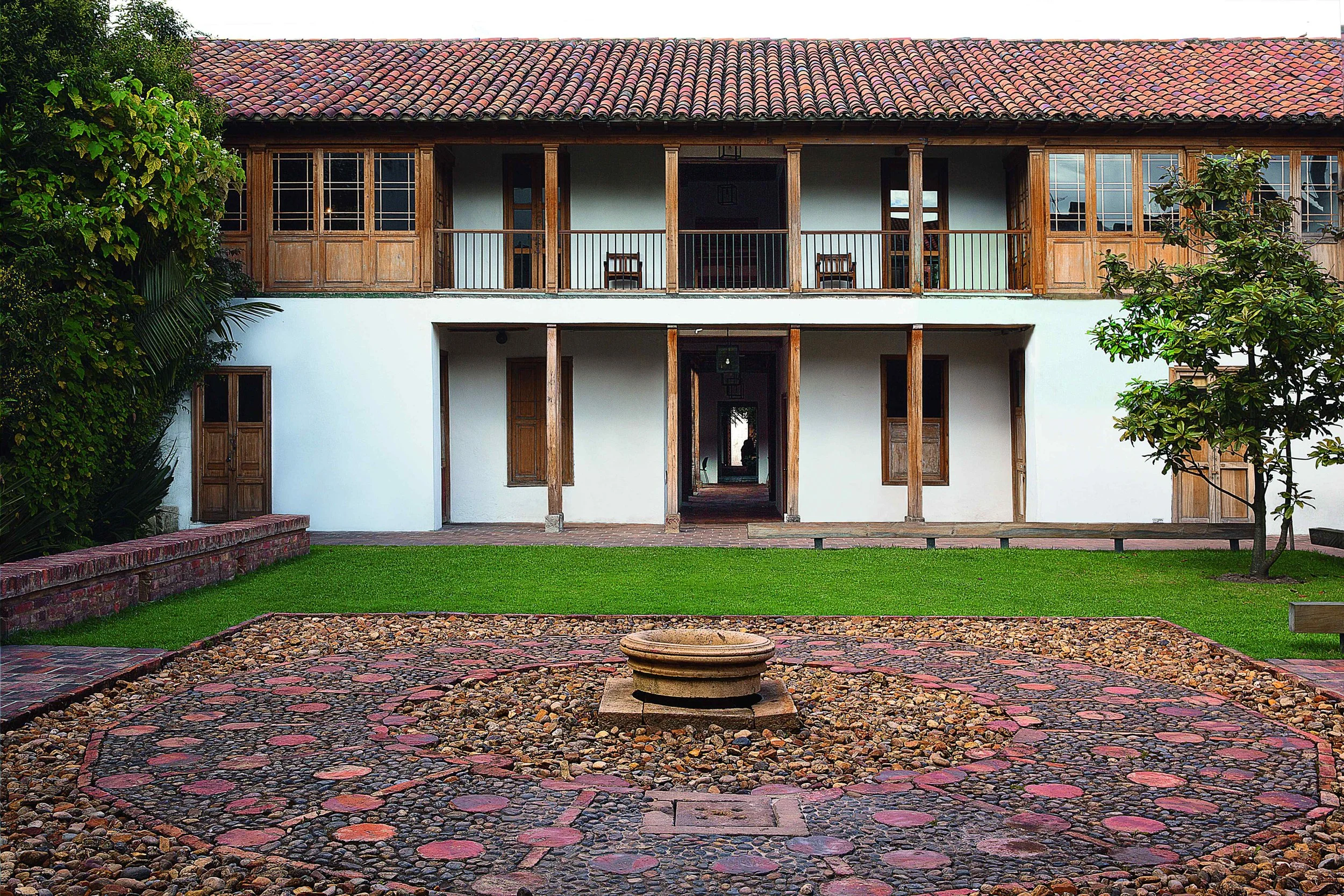 Two-story house with white walls, wooden window frames, and a red tiled roof, surrounded by greenery, with a stone fountain in the front yard.