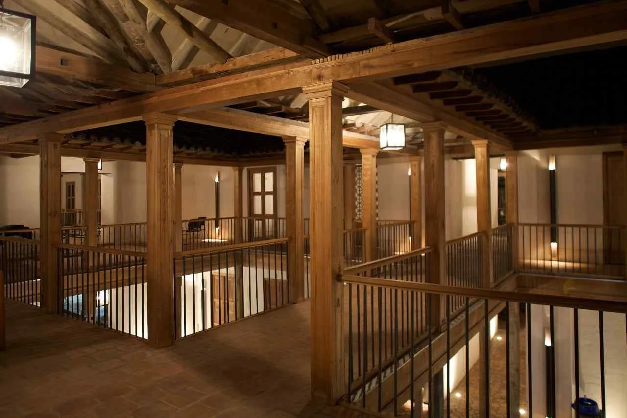 Interior of a building with wooden railings, columns, and a staircase, illuminated by wall-mounted lights and ceiling lights.