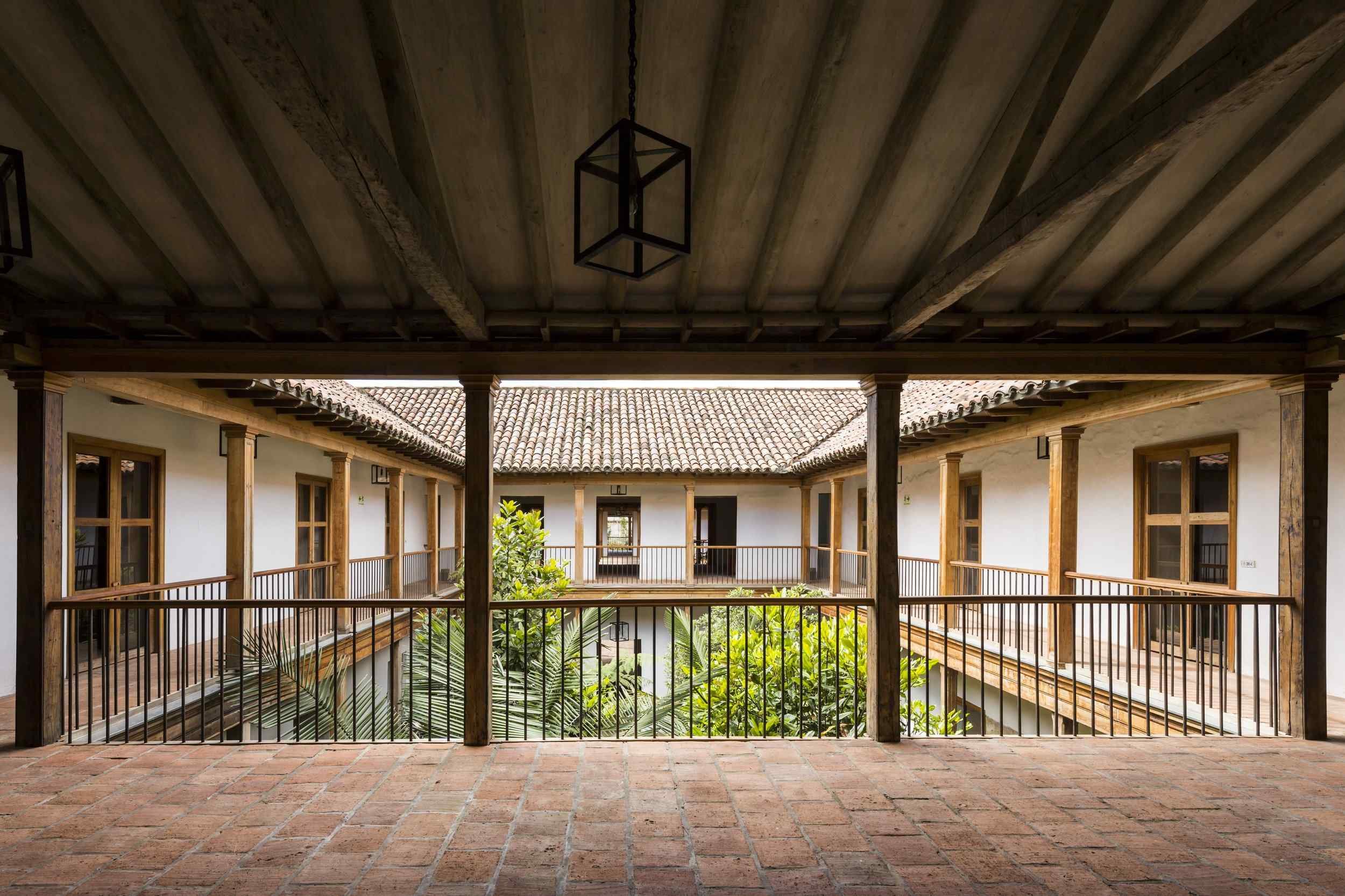 Interior view of a courtyard-style building with second-floor walkways, wooden railings, multiple doors and windows, tiled roof, and lush greenery in the center, viewed from a covered balcony.