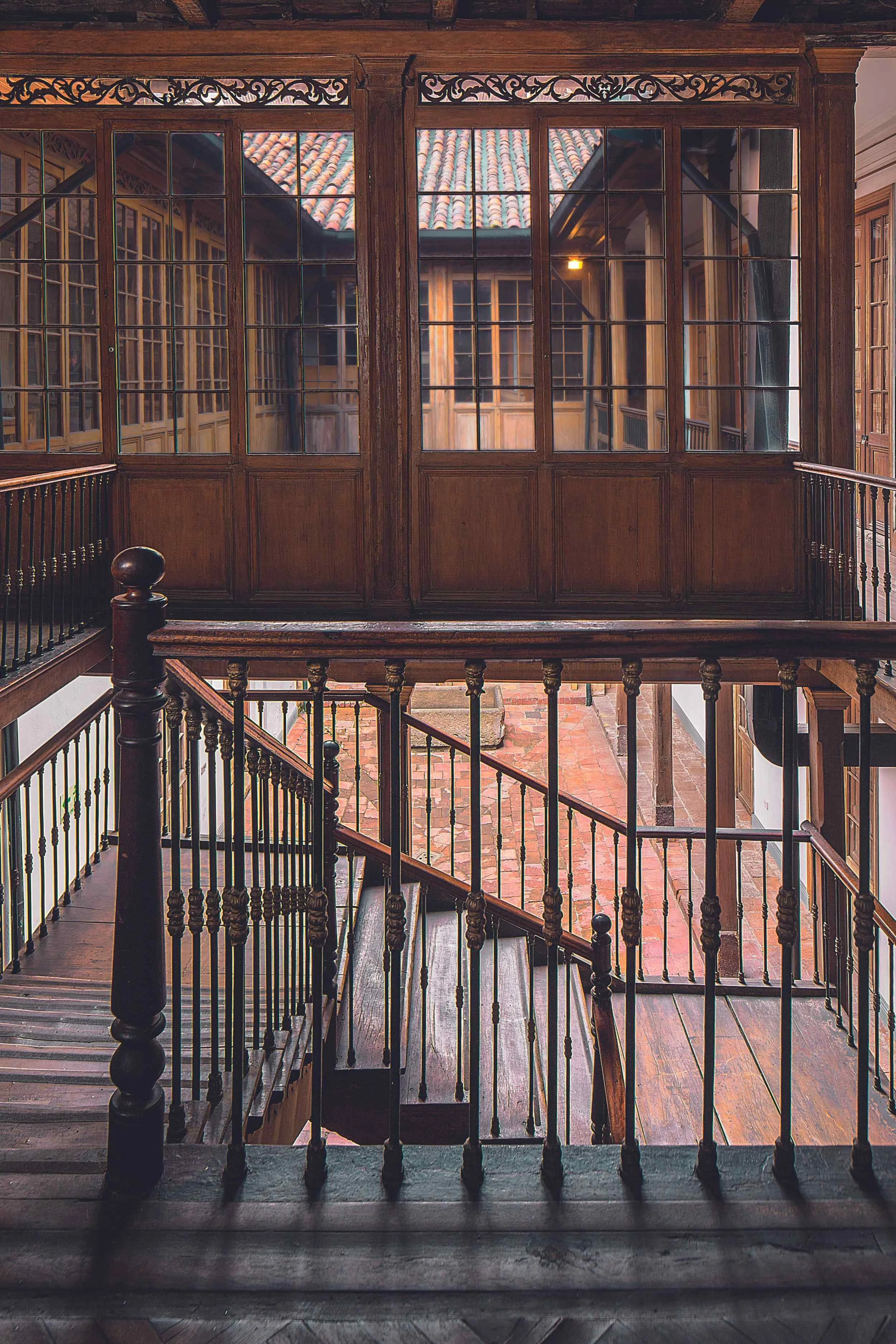 Interior view of a multi-level wooden staircase with ornate wooden railings, leading to a second-floor balcony with large windows and an indoor courtyard with brick flooring.
