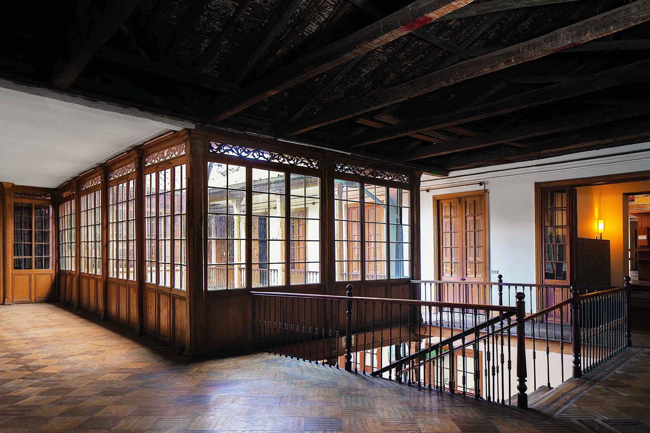 Interior view of a wooden staircase and a sunlit enclosed balcony with large glass-pane windows and wooden frames.