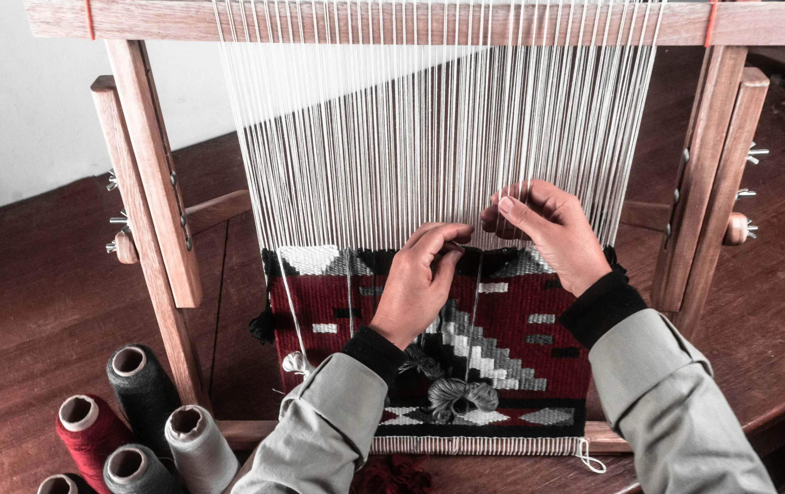 Person weaving fabric on a wooden loom, holding a weaving tool, with a piece of woven cloth on the loom.