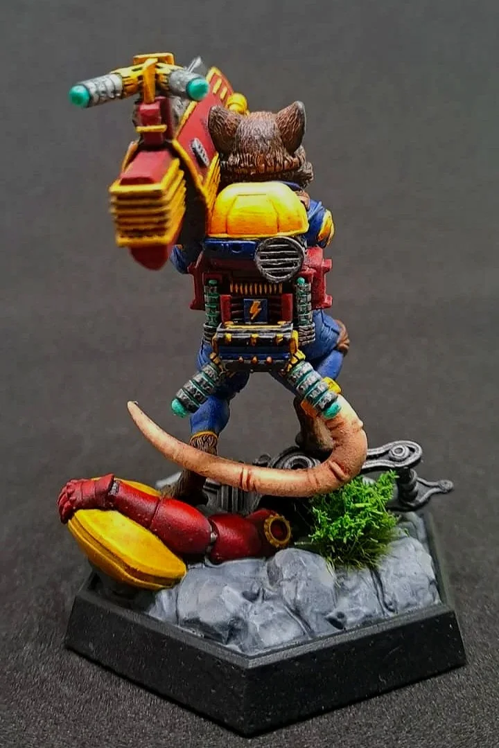 Details eines bemalten Spielzeug-Charakters, der ein Tier mit einer Rucksack-ähnlichen Vorrichtung auf dem Rücken trägt, auf einer quadratischen Basis mit Steinen, Pflanzen und technischen Elementen.