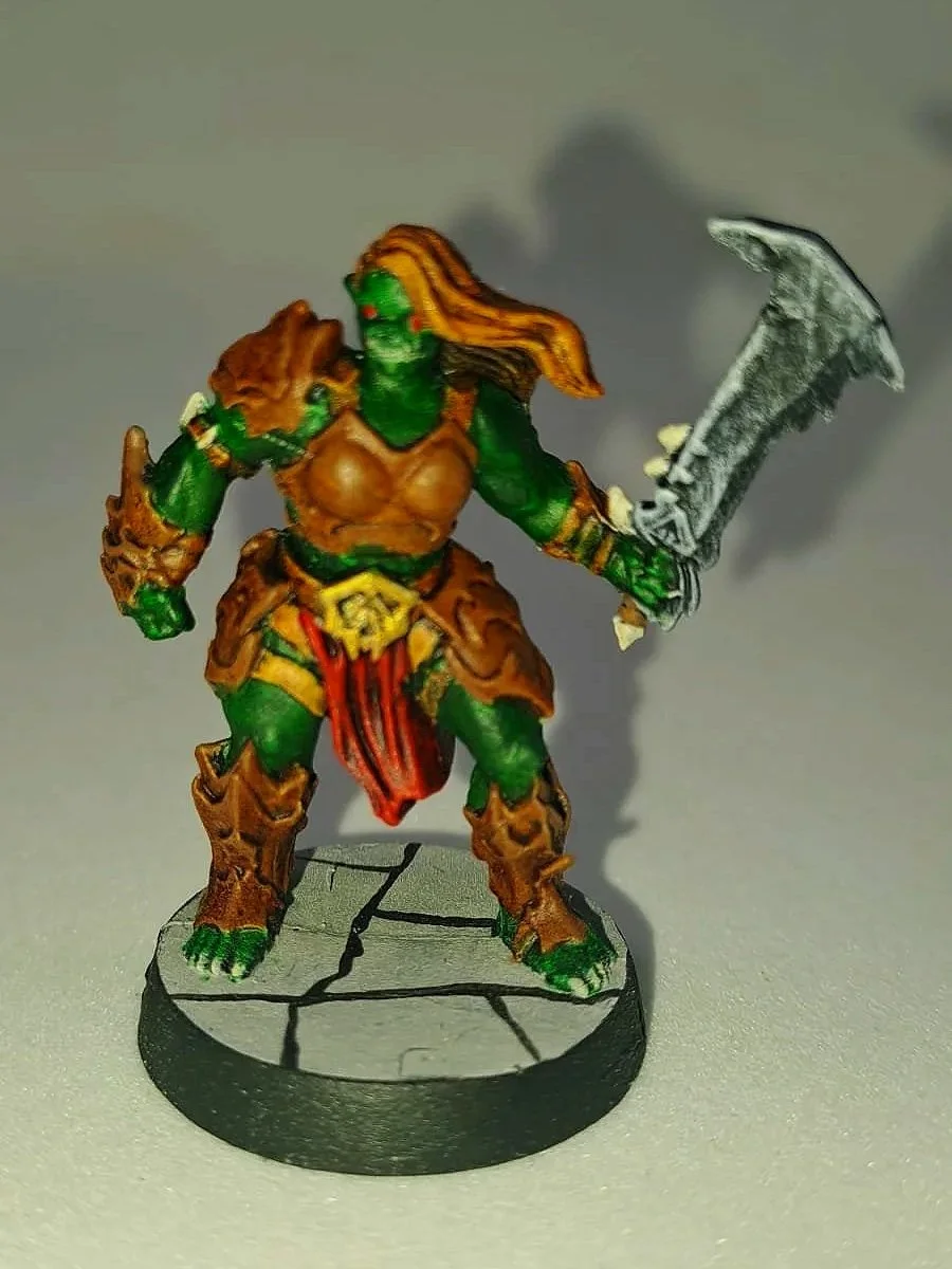Miniaturfigur eines grünen Ork-Kriegers mit brauner Rüstung, rot-schwarzem Kilt und großem Schwert auf einem runden Sockel