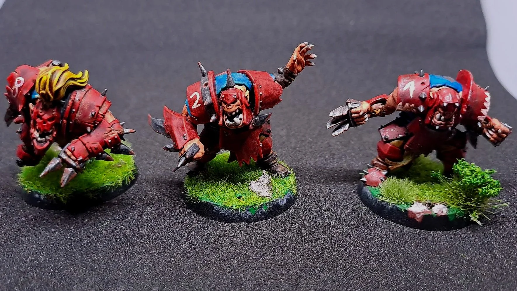 Drei detaillierte Figuren von Miniatur-Kriegern auf bases mit grüner Textur, bemalt in rot und schwarz mit Waffen und Helm-Details, im Fantasy-Stil.