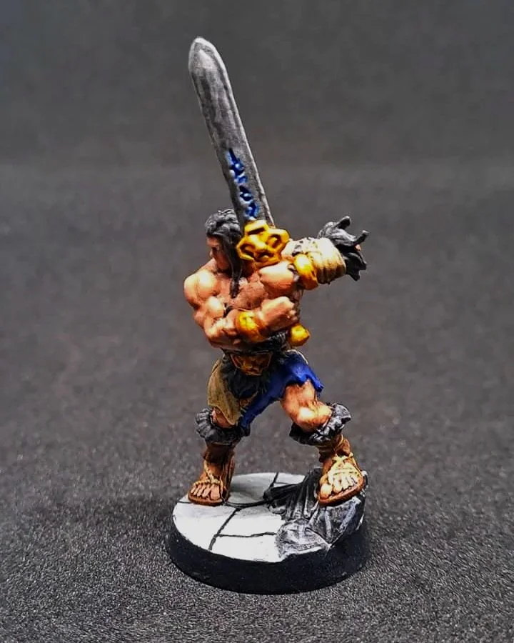Miniaturfigur eines muskulösen Kriegers mit einem großen Schwert, barfuß, in einer kampfbereiten Haltung auf einem runden Sockel.