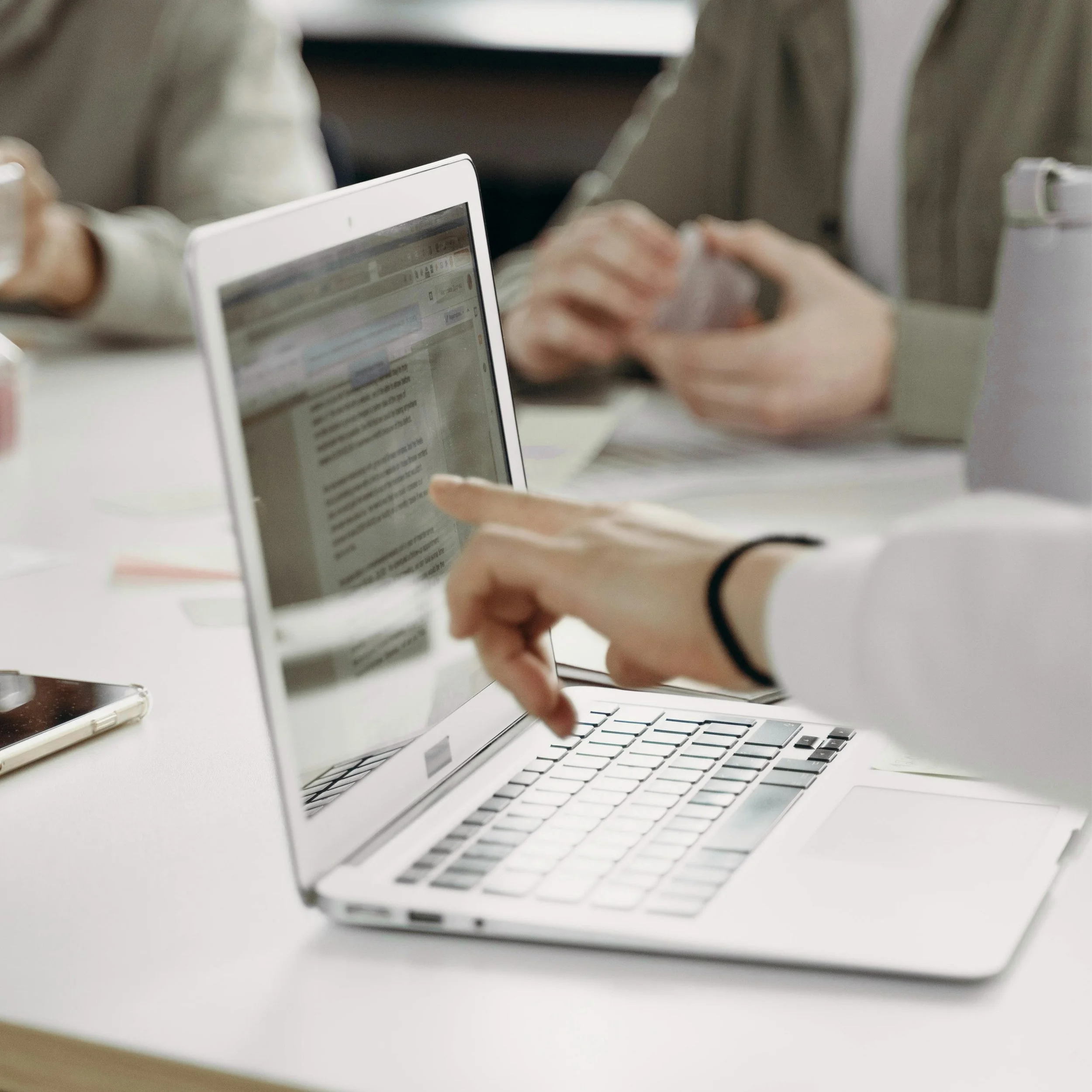 Une personne utilise un ordinateur portable, en tapant sur le clavier à l'aide de son index, dans un environnement de travail avec d'autres personnes assises autour de la table.