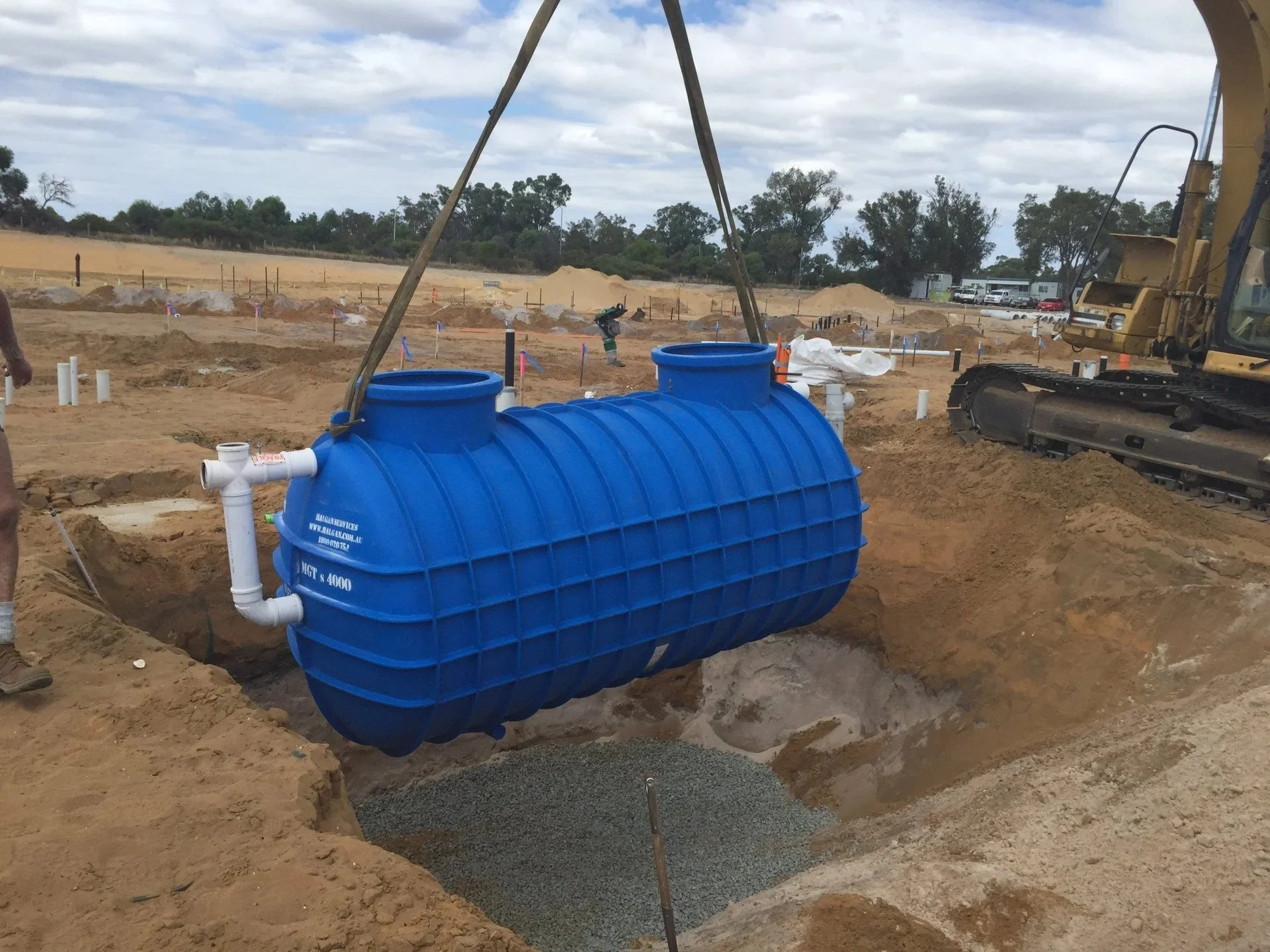 Blue underground septic tank being lowered by crane into a trench on a construction site with excavator nearby and cloudy sky overhead.