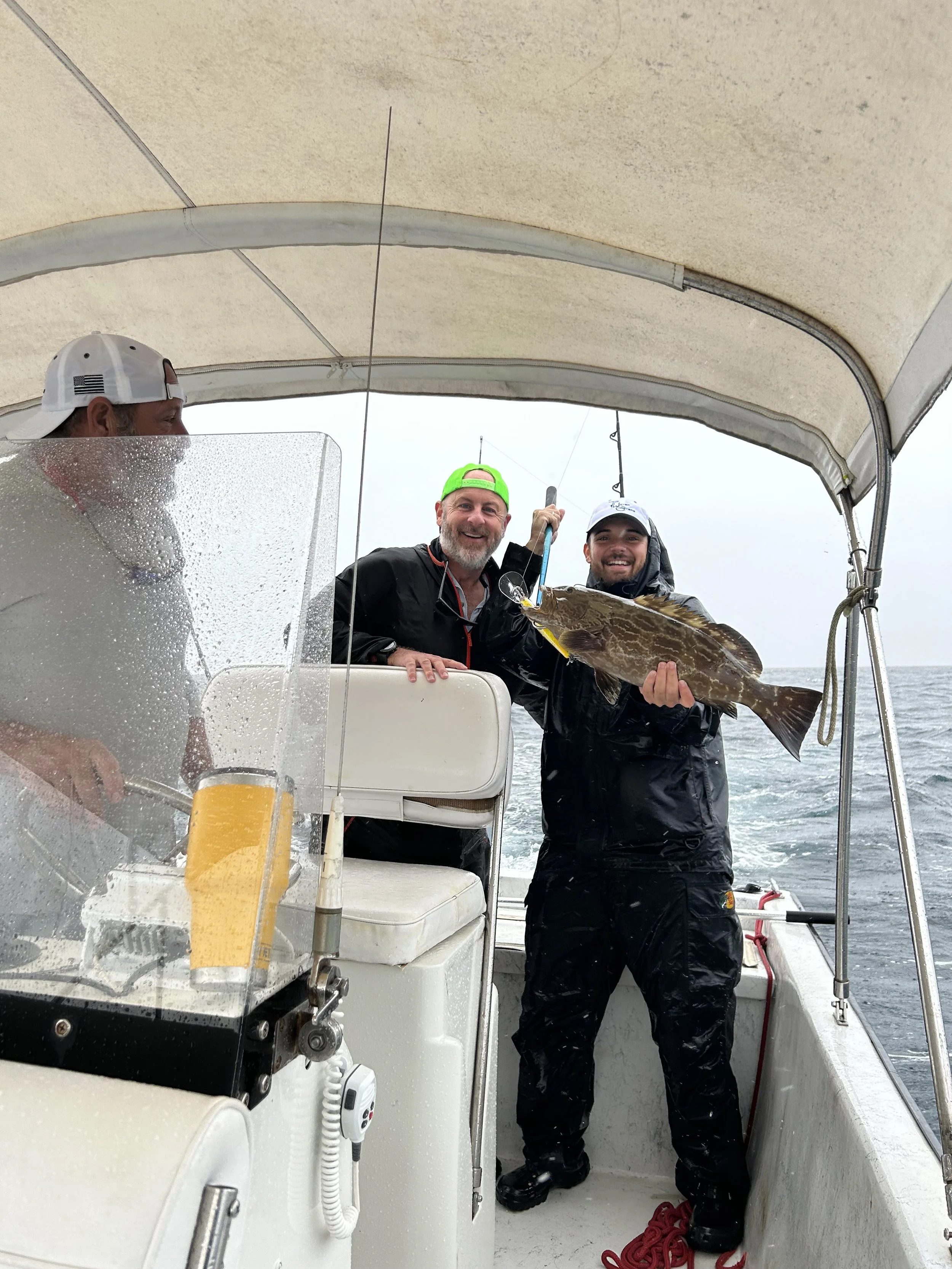 Three men on a boat, with one holding a large fish, celebrating a successful catch during a fishing trip on a cloudy day at sea.