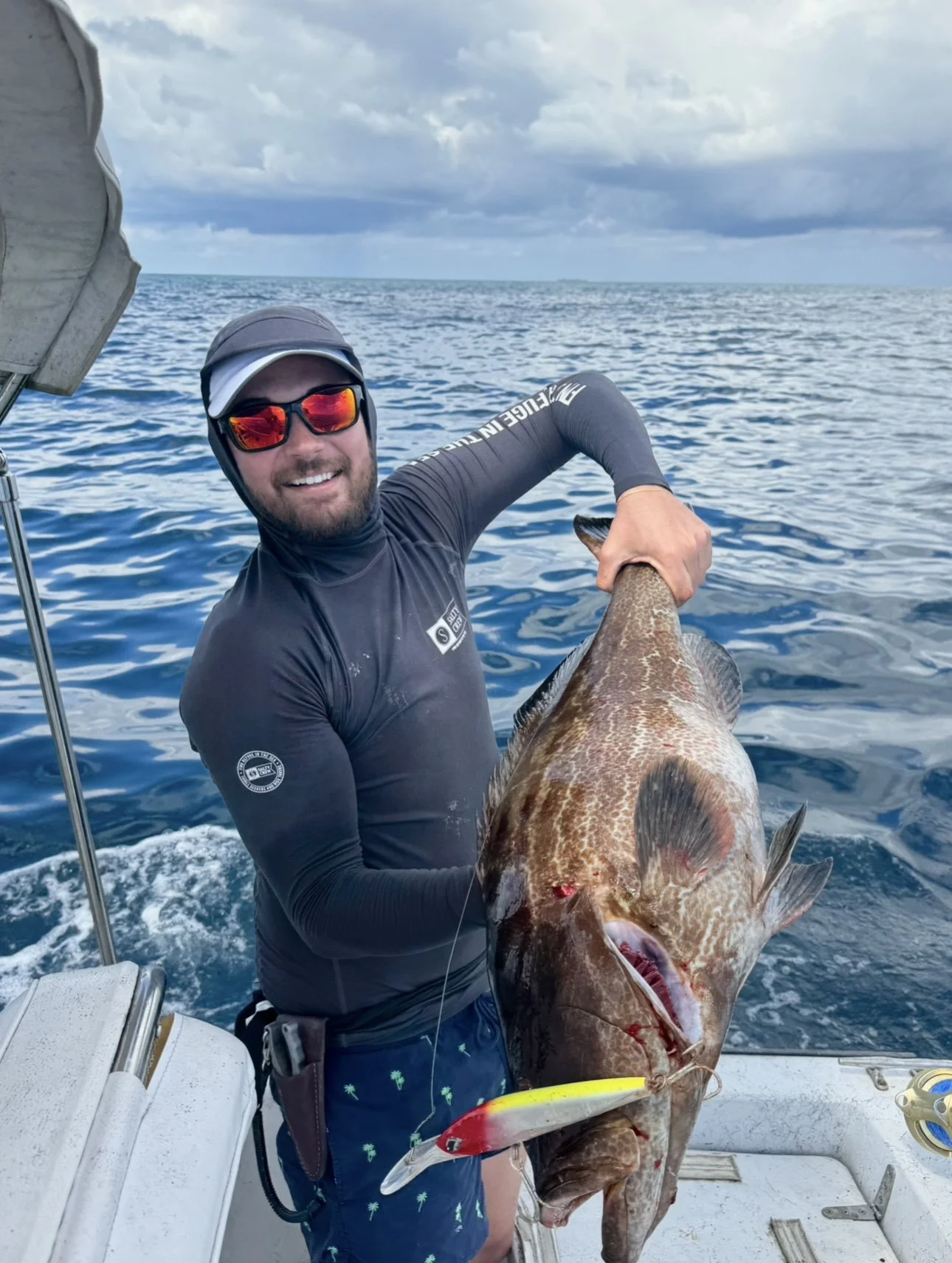 Man holding a large fish on a boat out at sea, cloudy sky, wearing sunglasses, a long-sleeve shirt, and swim trunks.