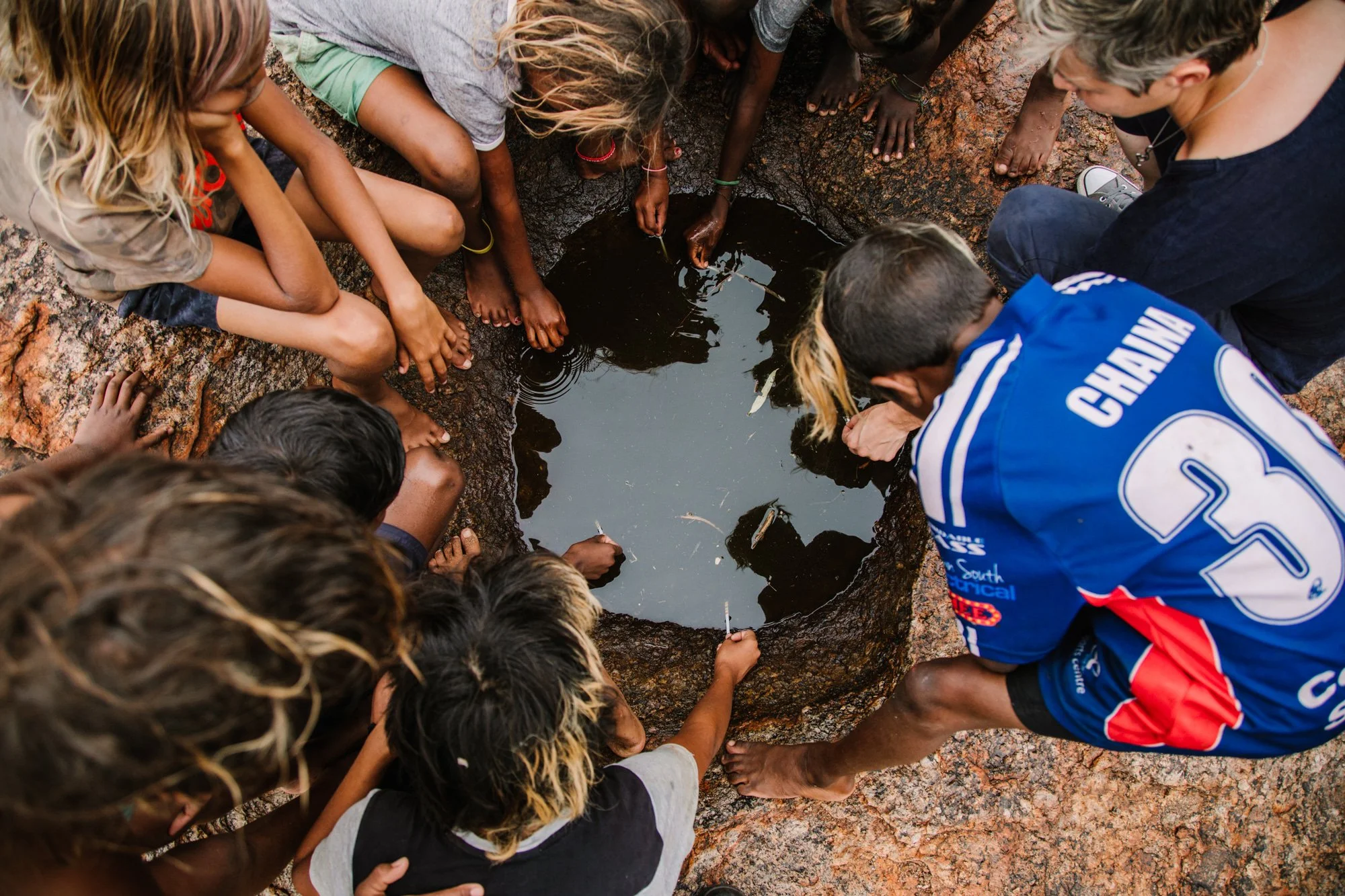 A group of children and an adult gathered around a small waterhole on rocky ground, with some children holding small sticks. Photographed on APY Lands with Mimili Anangu School for Department of Education South Australia