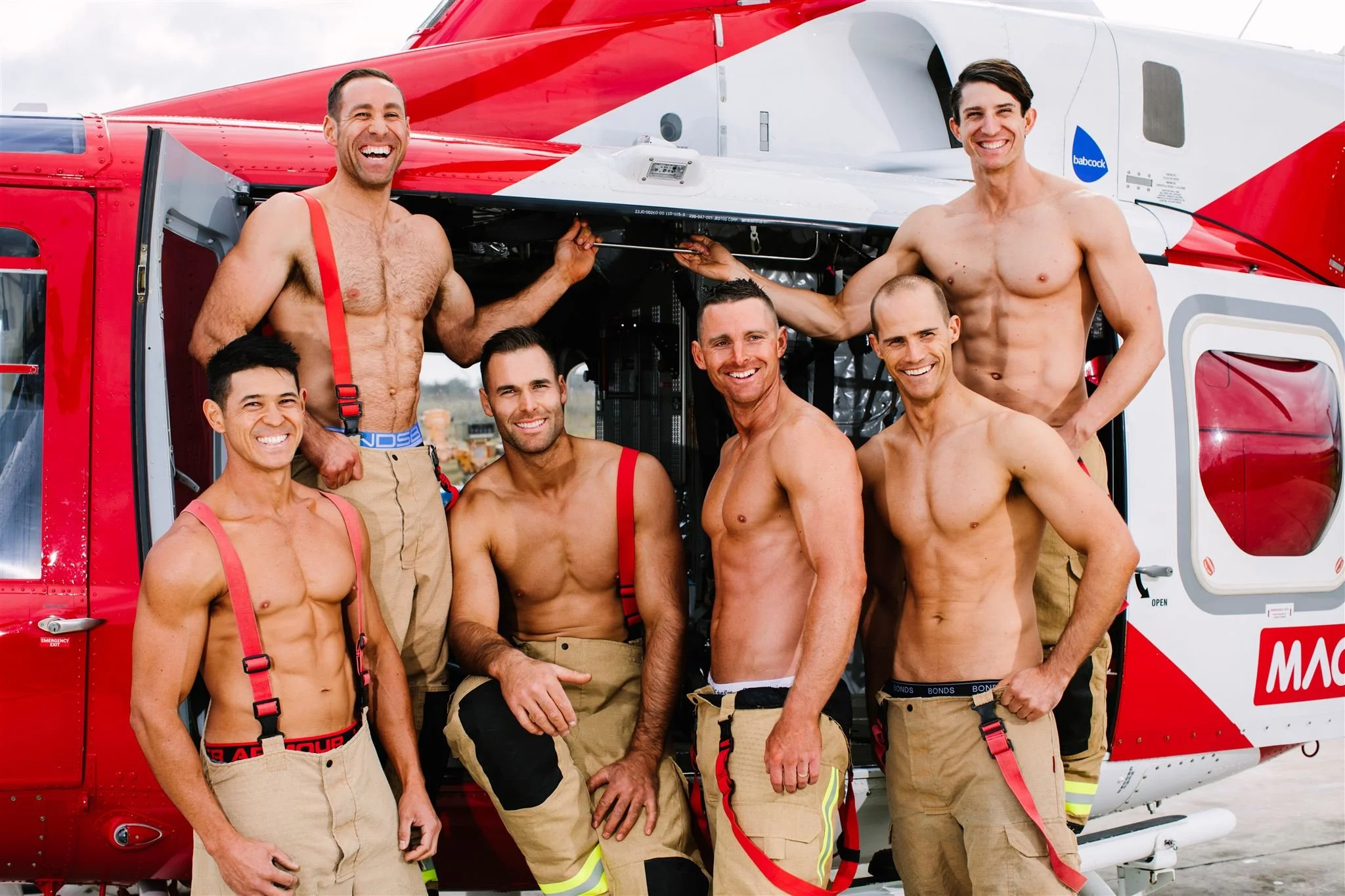 Group of seven shirtless firefighters in front of a red and white rescue helicopter, smiling and posing for the photo.