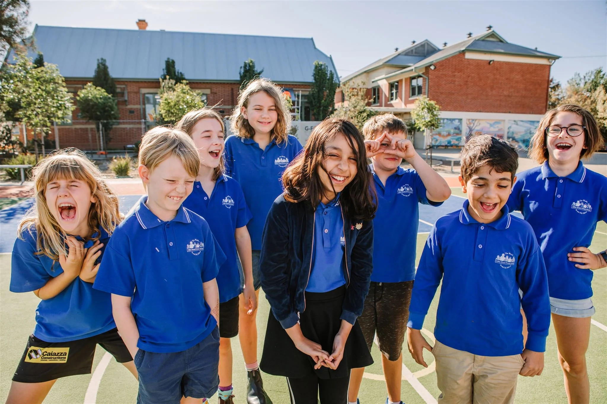 A group of eight children from Gilles Street Primary School Adelaide and Windmill Theatre Company Creation Creation in blue shirts standing together on a school playground, all laughing and smiling.