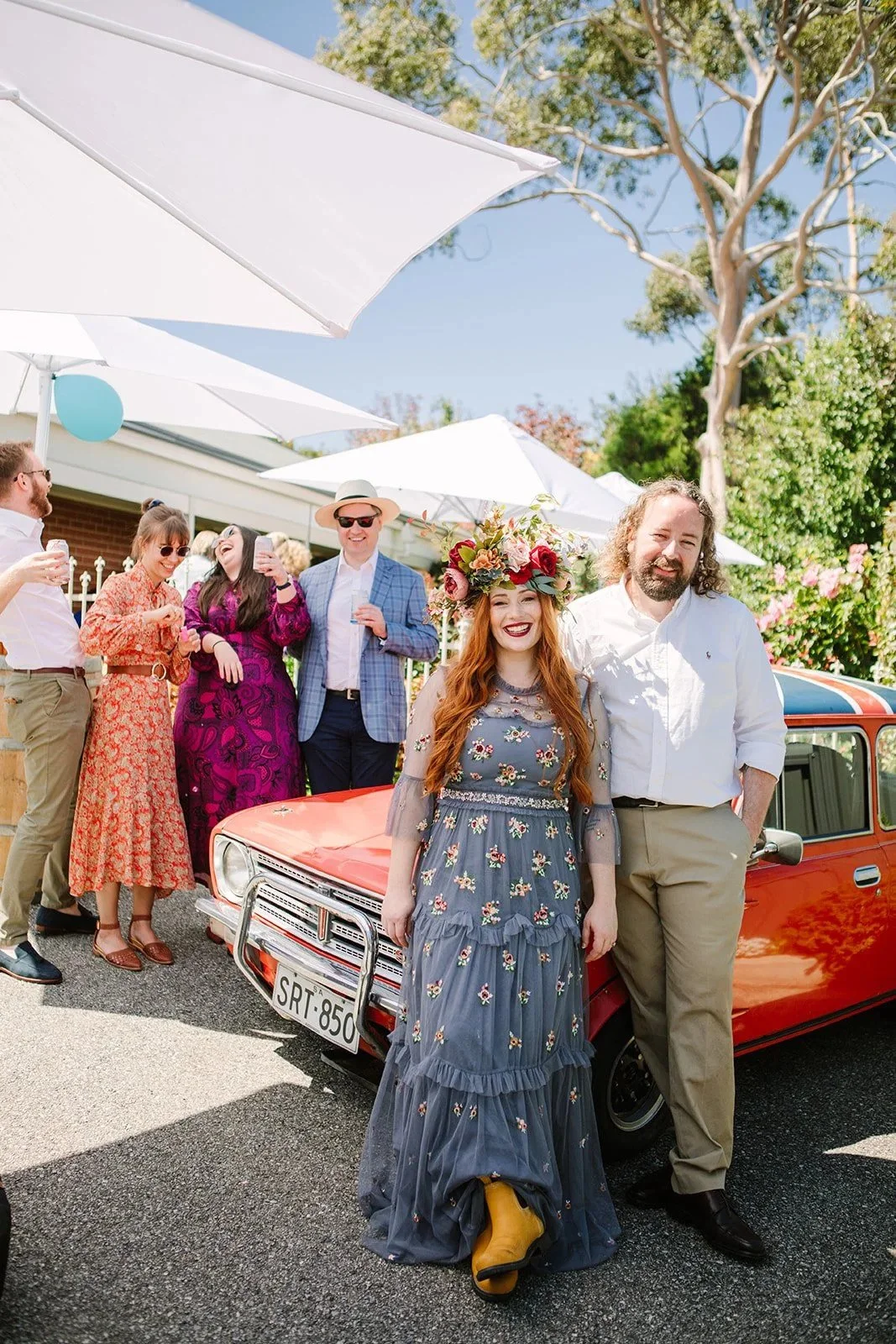 Group of friends celebrating outdoors under white umbrellas, with two women in the foreground, one wearing a floral dress with a flower crown and the other in a purple dress, standing next to a red vintage car, smiling and enjoying the sunny day.