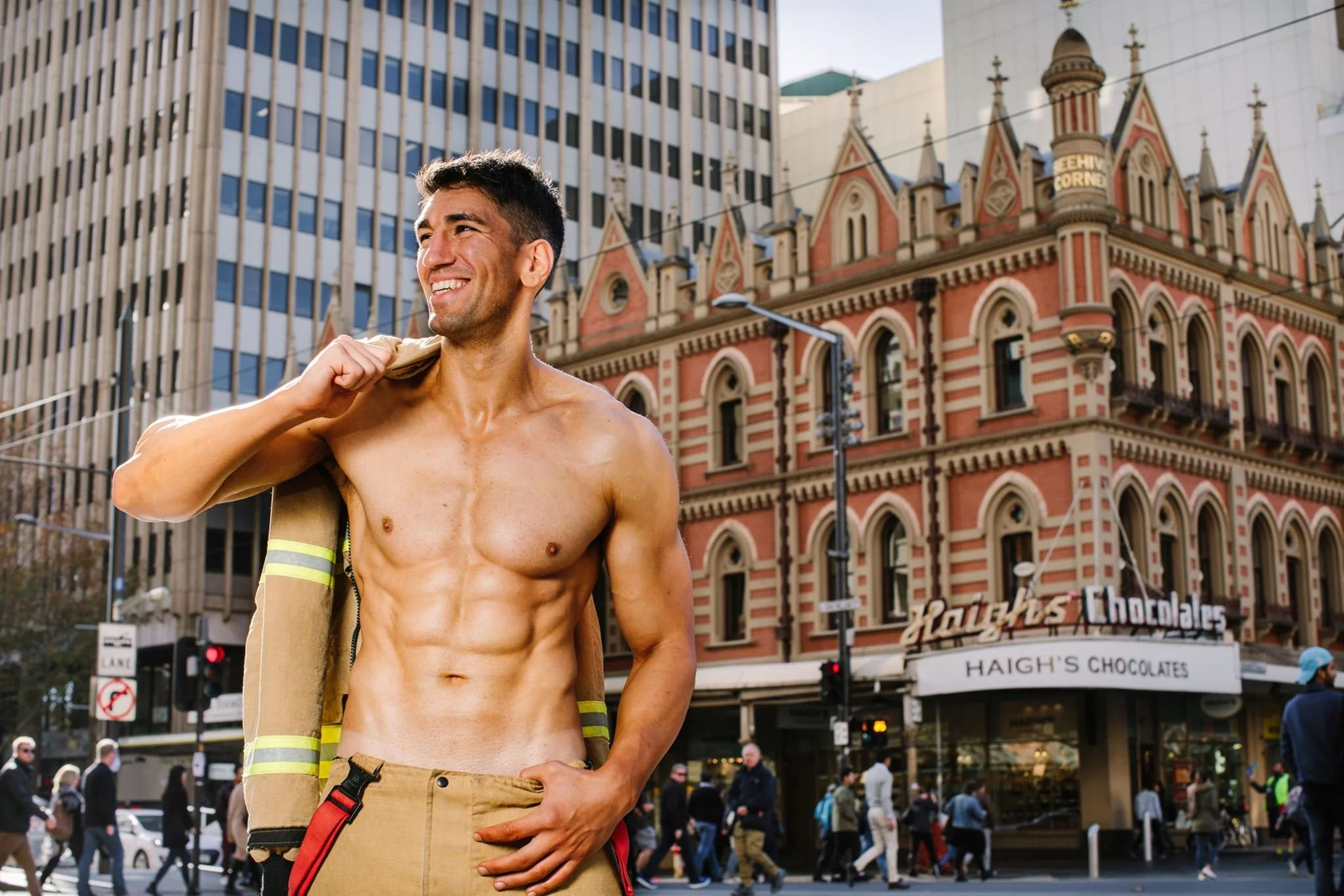 A shirtless firefighter carrying his coat over his shoulder, standing on a city street with tall buildings and pedestrians in the background, the location is Rundle Mall Beehive Corner, Adelaide, South Australian