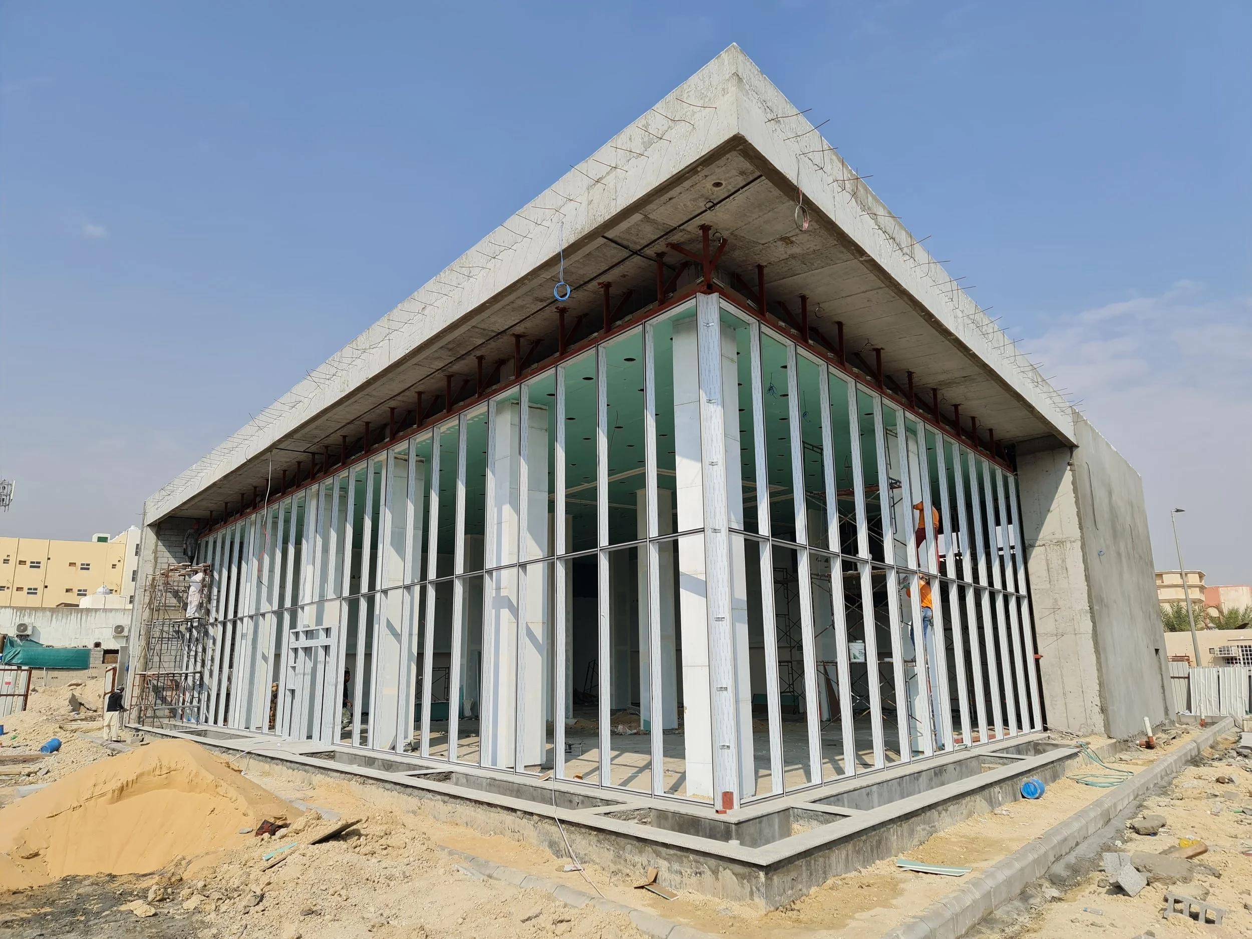 A modern building under construction with steel framing and large glass windows, surrounded by construction debris and sand, with a blue sky in the background.
