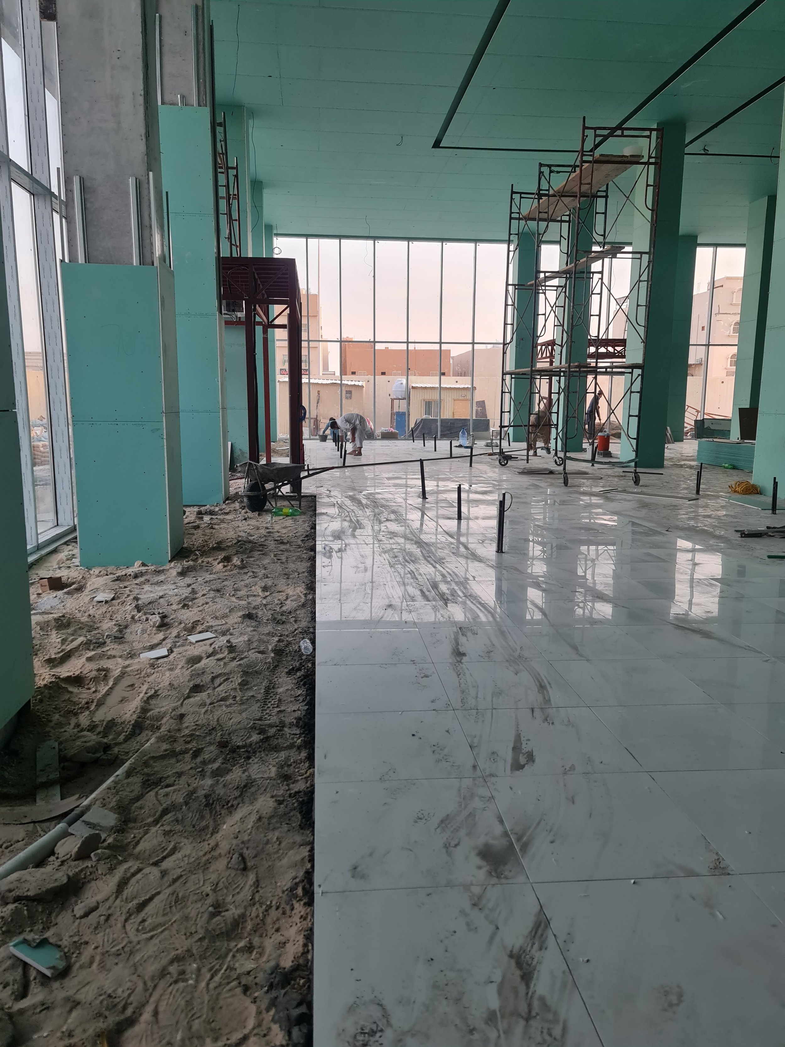 Interior of a building under construction with large glass windows, scaffolding, construction workers, and a partially finished shiny tile floor.