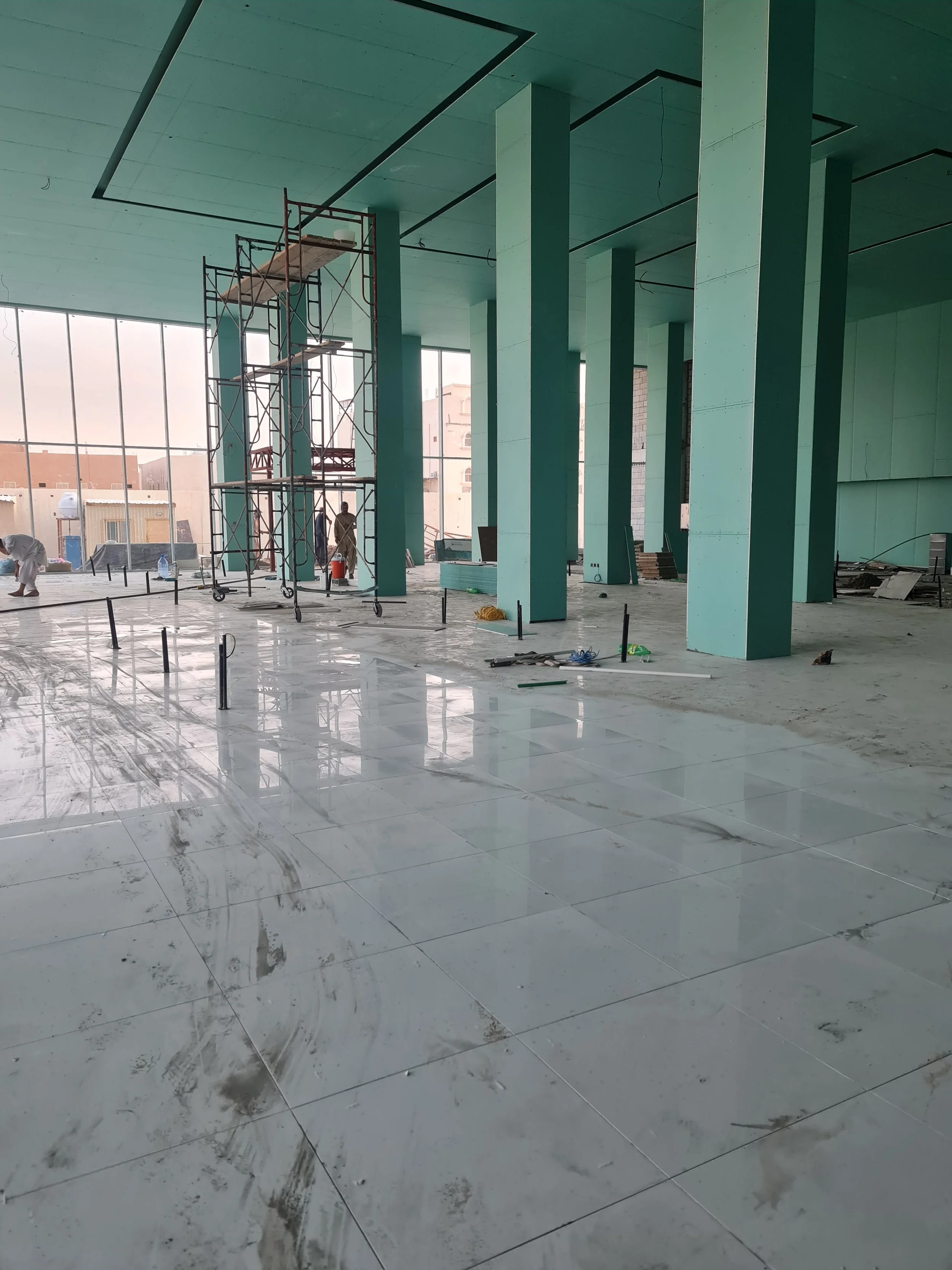 Interior of a building under construction with large windows, green columns, and workers installing floor tiles.