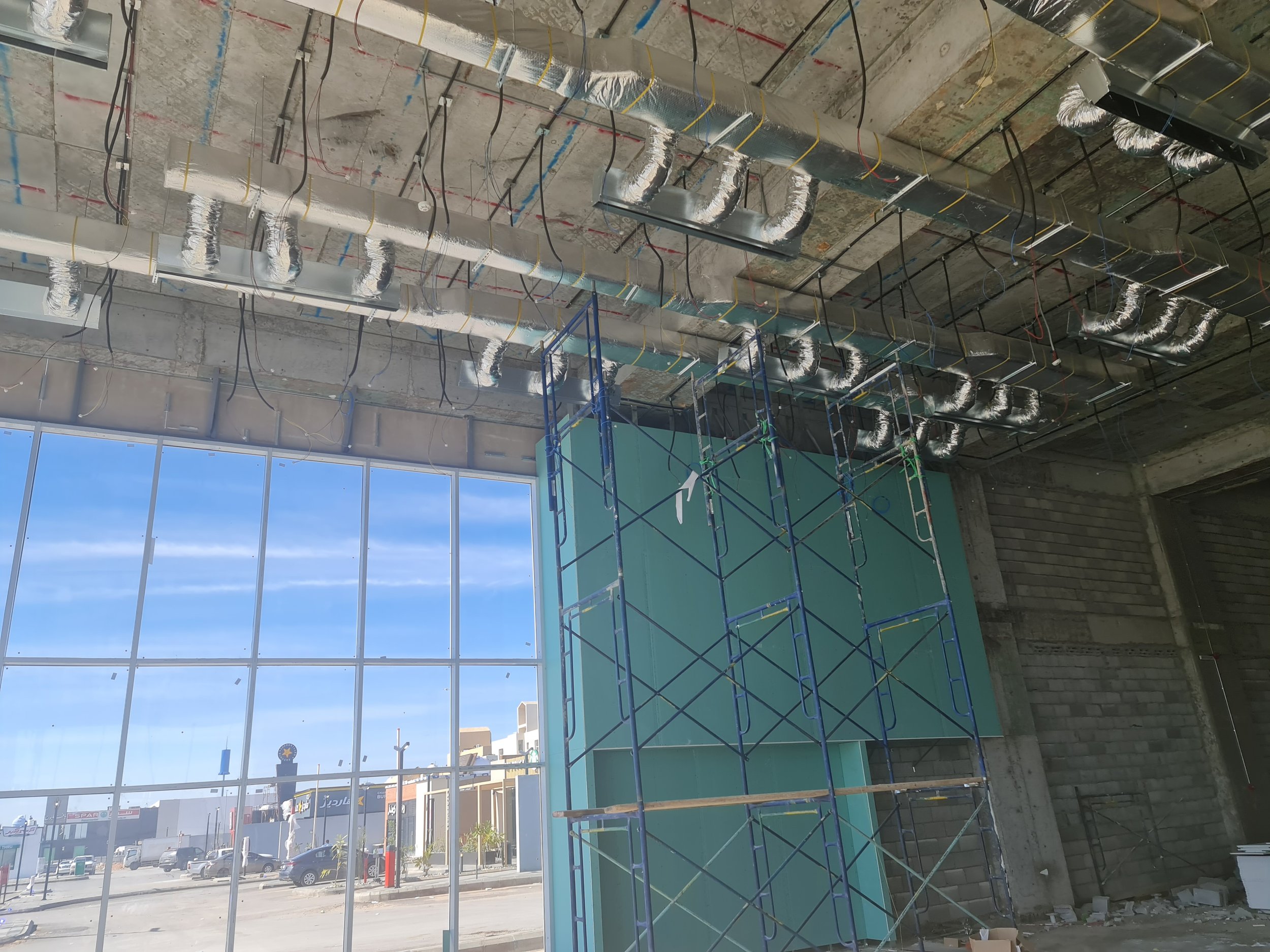 Construction interior with exposed ductwork, scaffolding, and a large window showing an exterior parking lot and buildings.