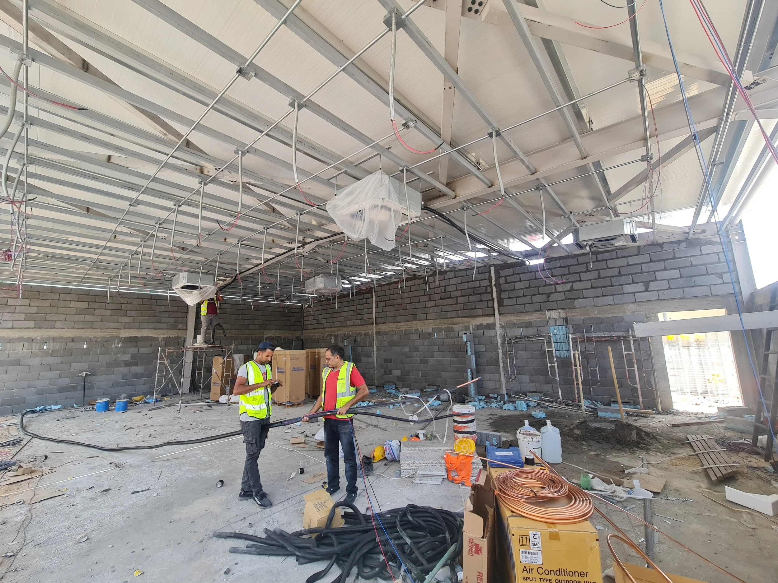 Construction workers inside a building under construction, working on electrical wiring and other installation tasks, with building materials and tools scattered around.