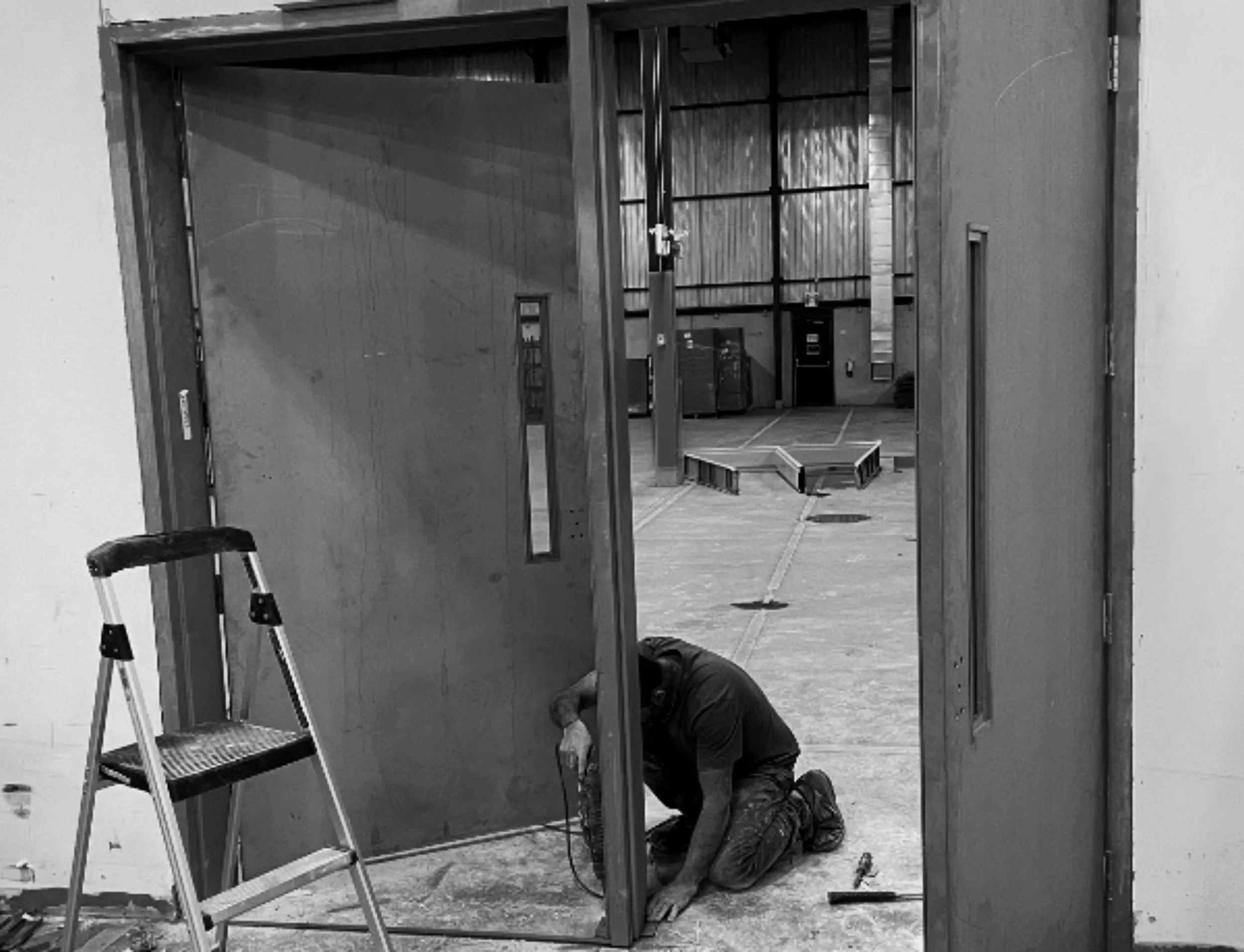 A worker kneeling on the floor installing a large sliding door in a warehouse or workshop. The door is partially open, revealing more equipment and space inside. A ladder is placed to the left of the worker.