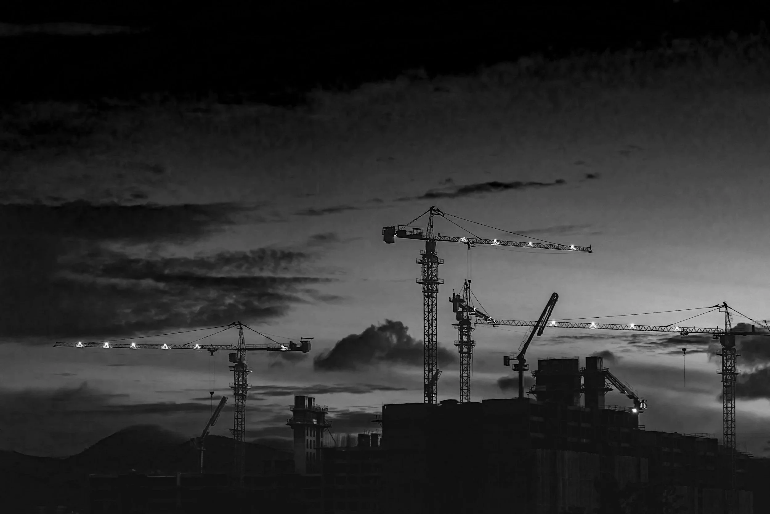Black and white photo of a construction site with multiple tall cranes and building structures against a cloudy sky at dusk.