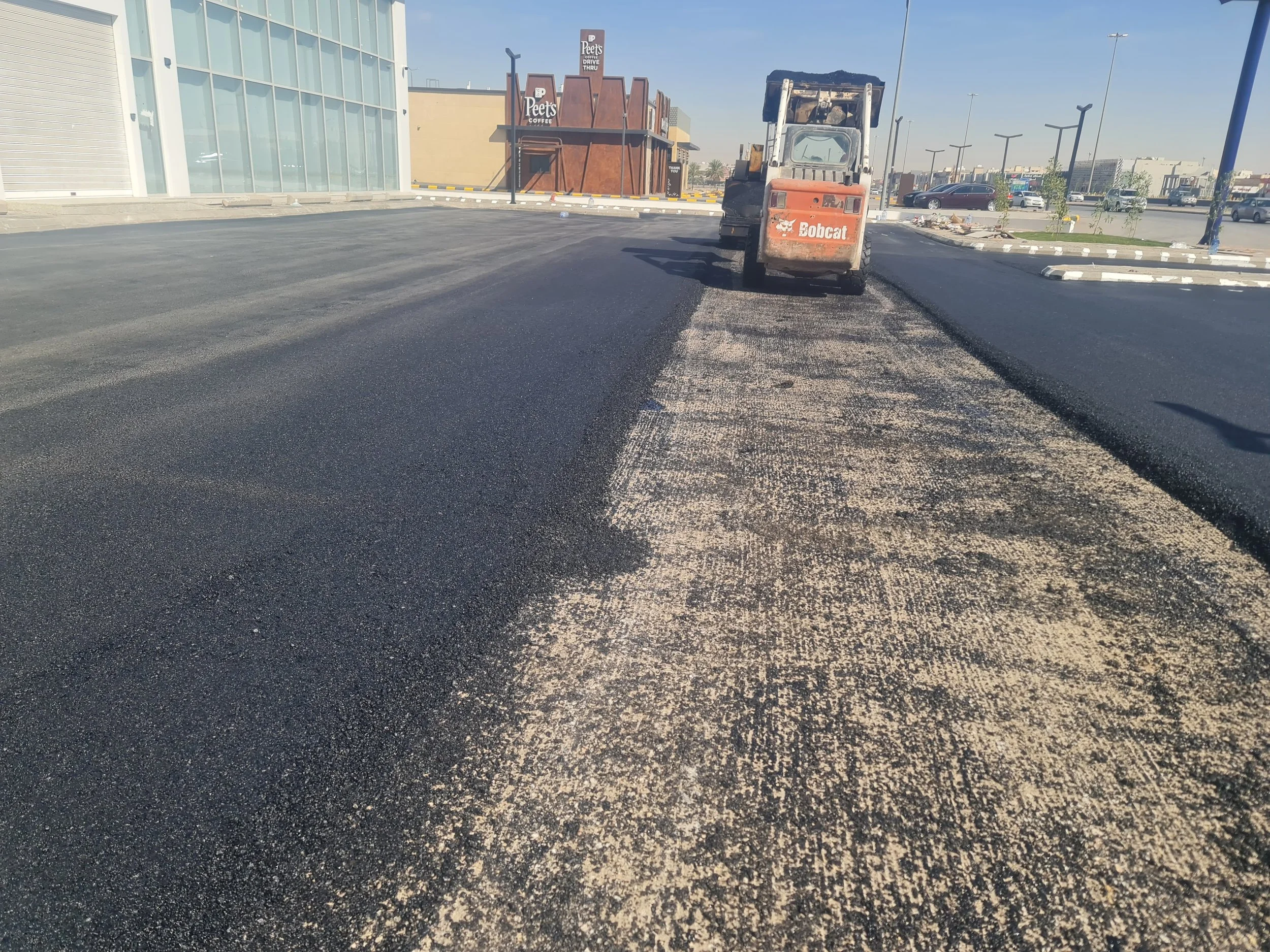 Construction site showing a road being paved with asphalt, with a compacting machine on the right, and a commercial building in the background.