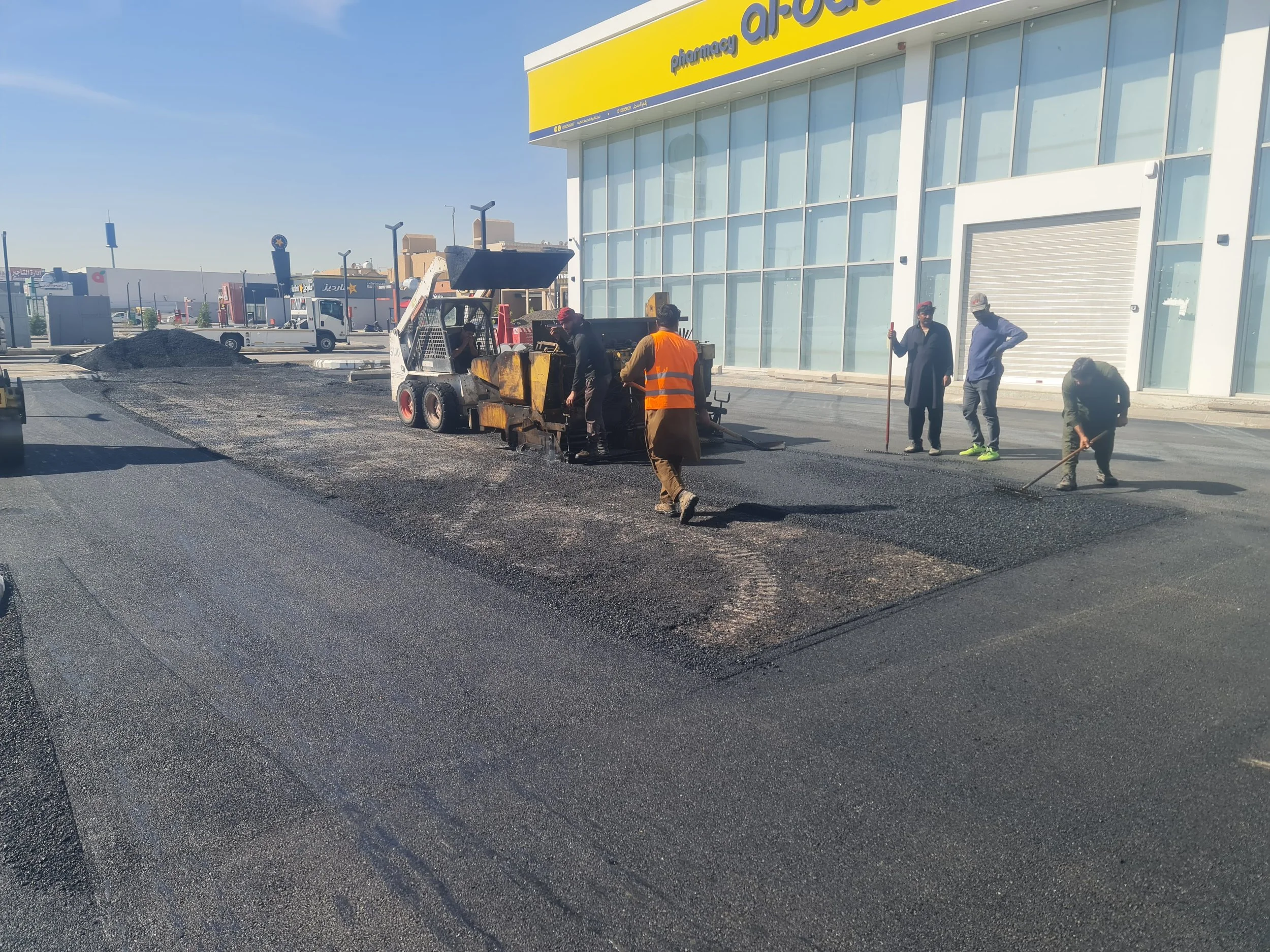 Workers paving a parking lot in front of a store with a yellow sign, using machinery and tools to spread asphalt.