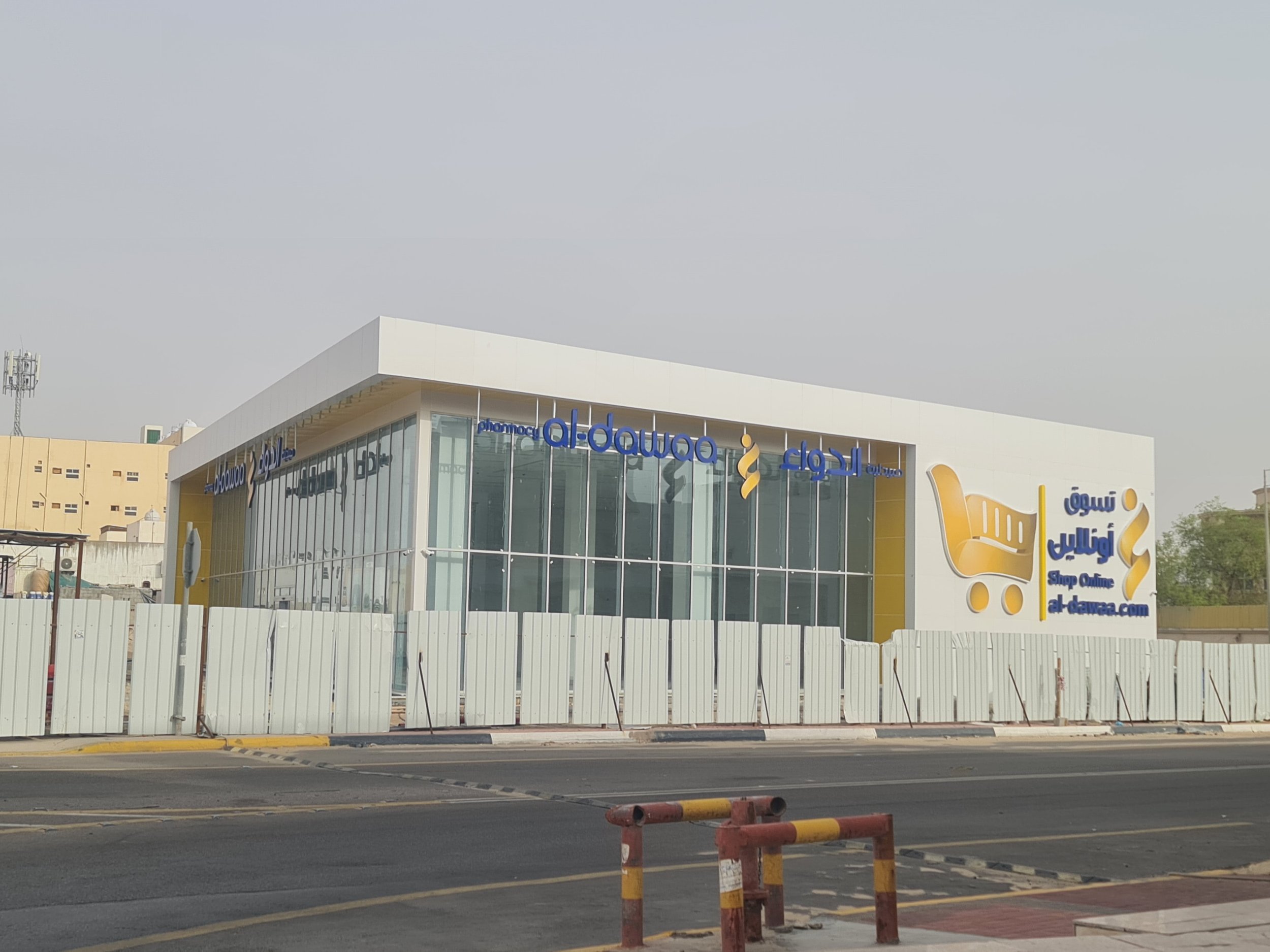 Outside view of a modern building with large glass windows, featuring signs in Arabic and English for an online shopping site al-dawaa.com, with a yellow shopping cart logo on the right side.