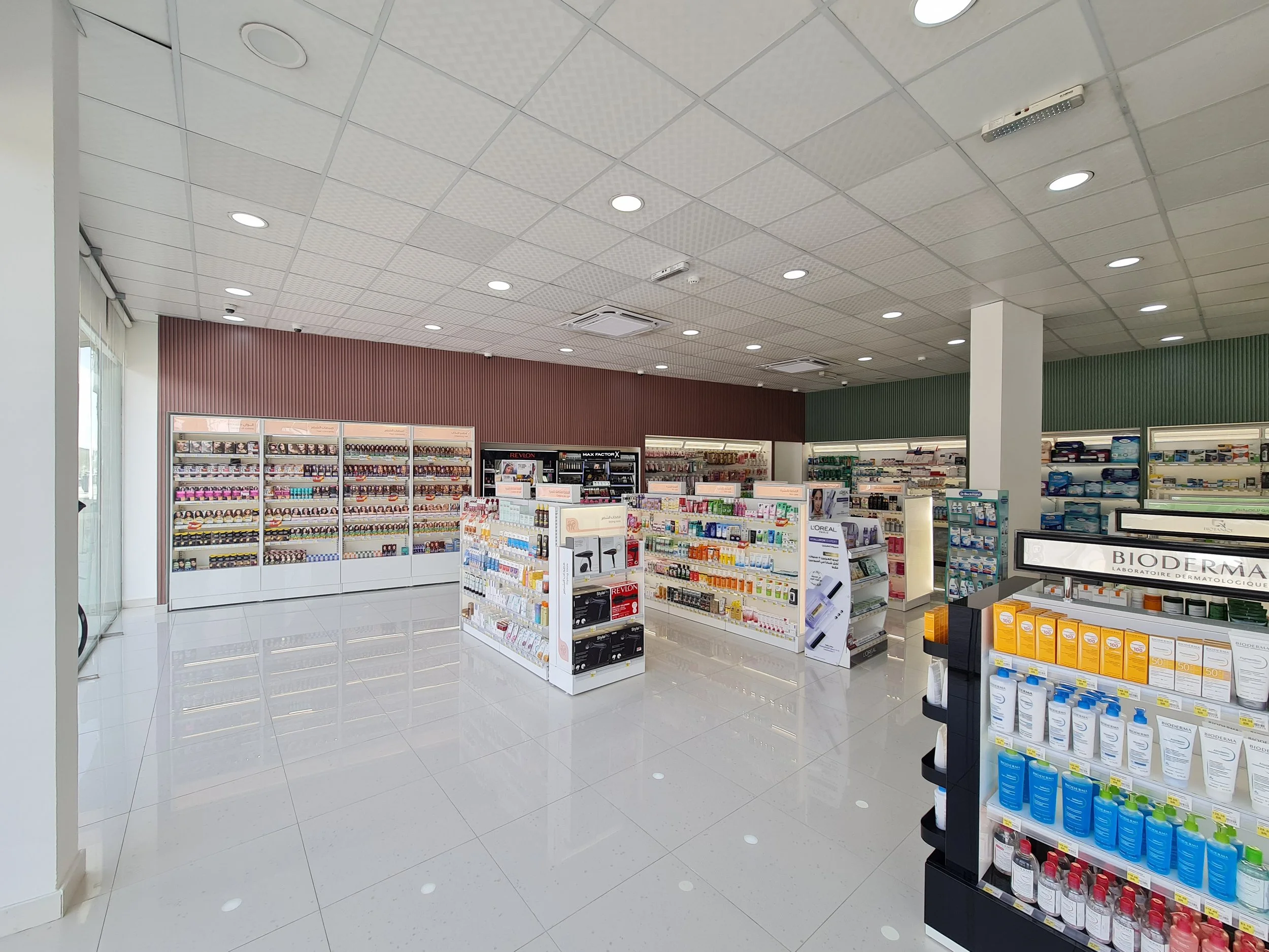 Inside a pharmacy with shelves stocked with various skincare, makeup, and health products, including a display for Bioderma, and a large window on the left side bringing natural light.