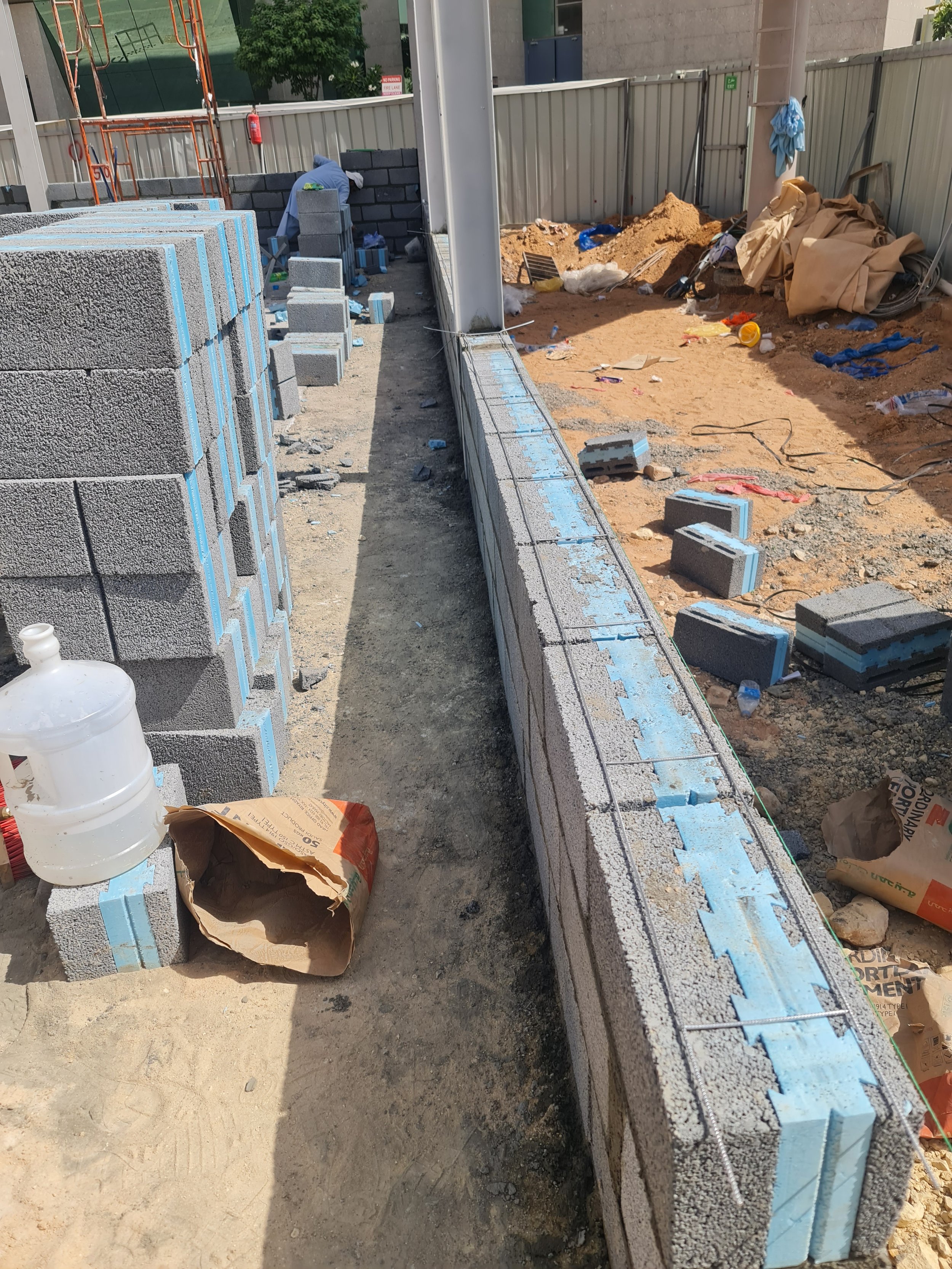 Construction site with stacks of concrete blocks, a worker laying blocks, and construction tools scattered around.