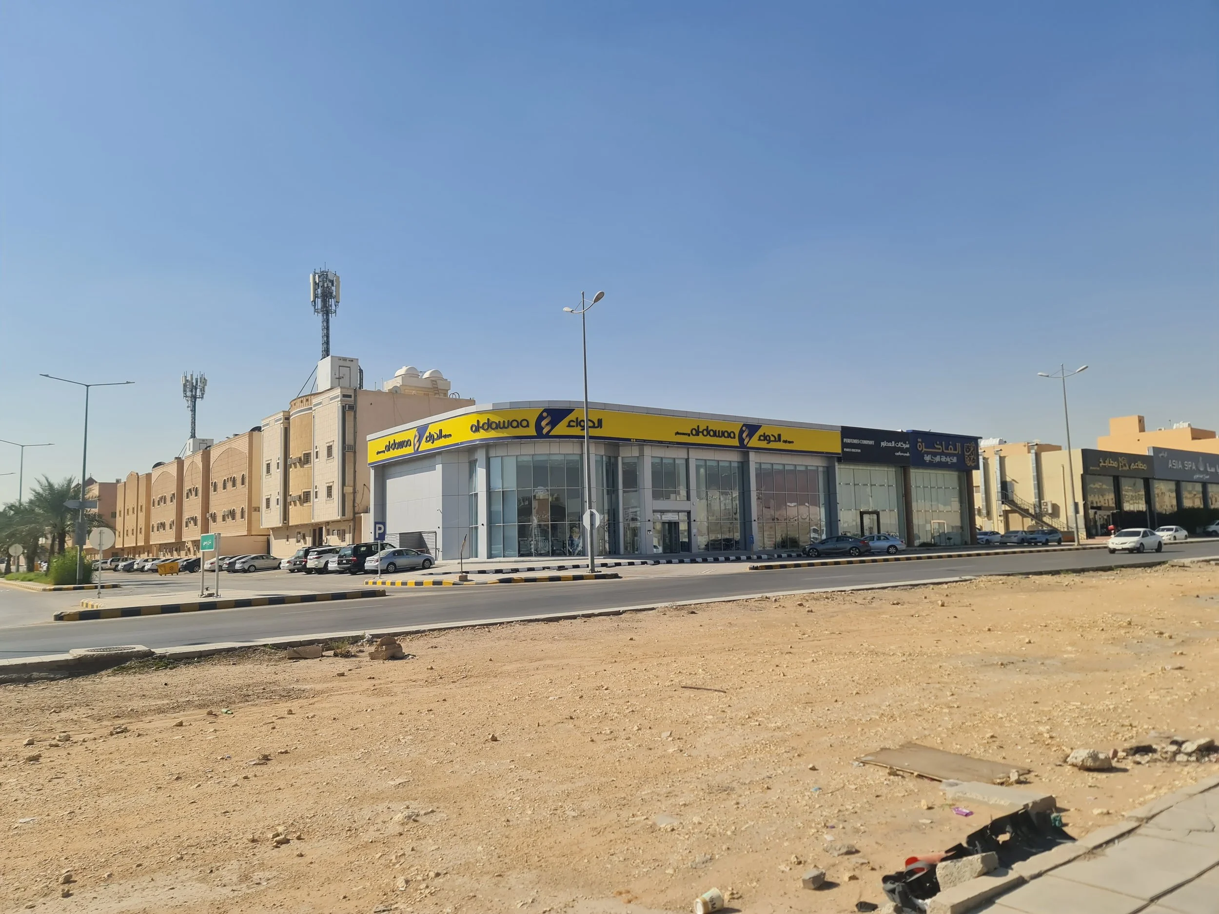 Commercial building with glass fronts and yellow signs that read 'aldawaa,' along with other business signs in Arabic, located on a street with multiple parked cars and street lamps under a clear blue sky.