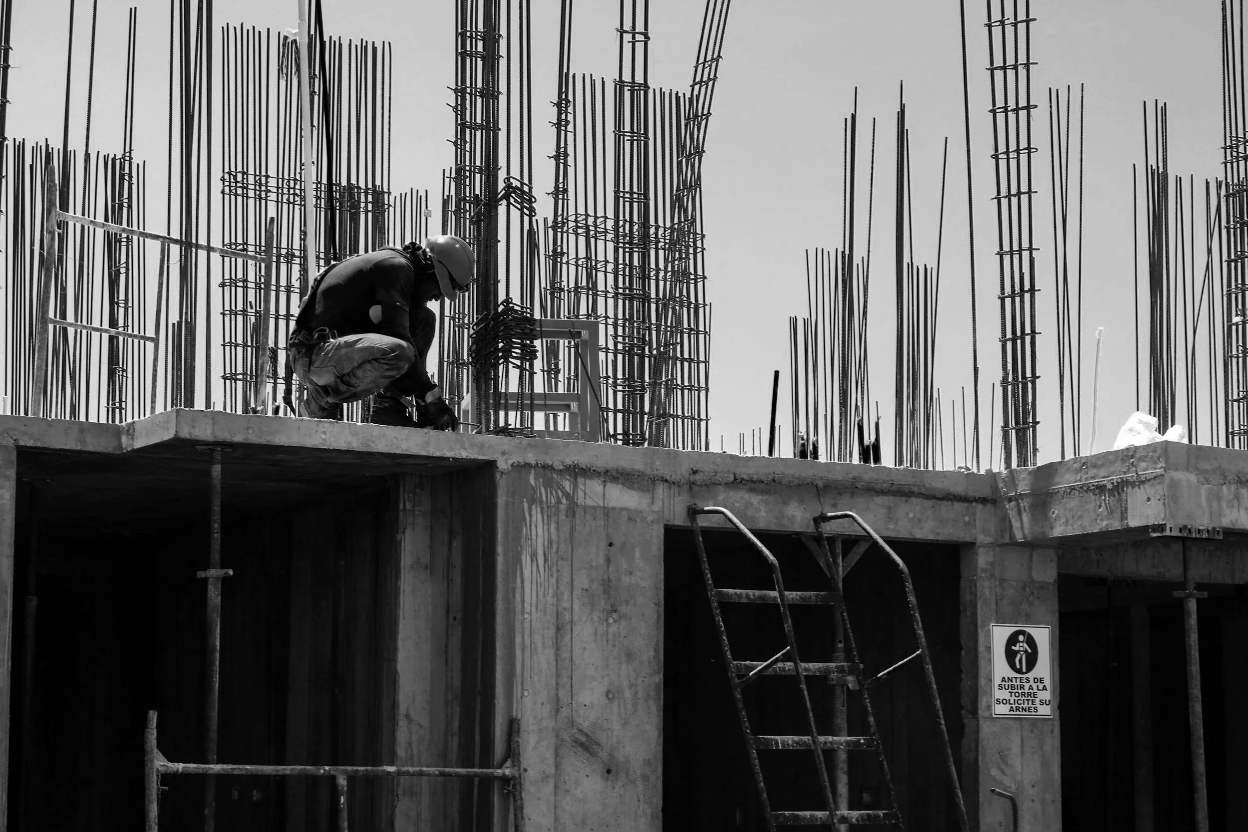A construction worker wearing a safety helmet kneels on a concrete ledge, working on rebar at a building site. Steel rebar protrudes vertically around the worker, and a staircase ladder is leaning against the concrete structure. There is a safety sign on the wall instructing to ask for permission before climbing the tower.