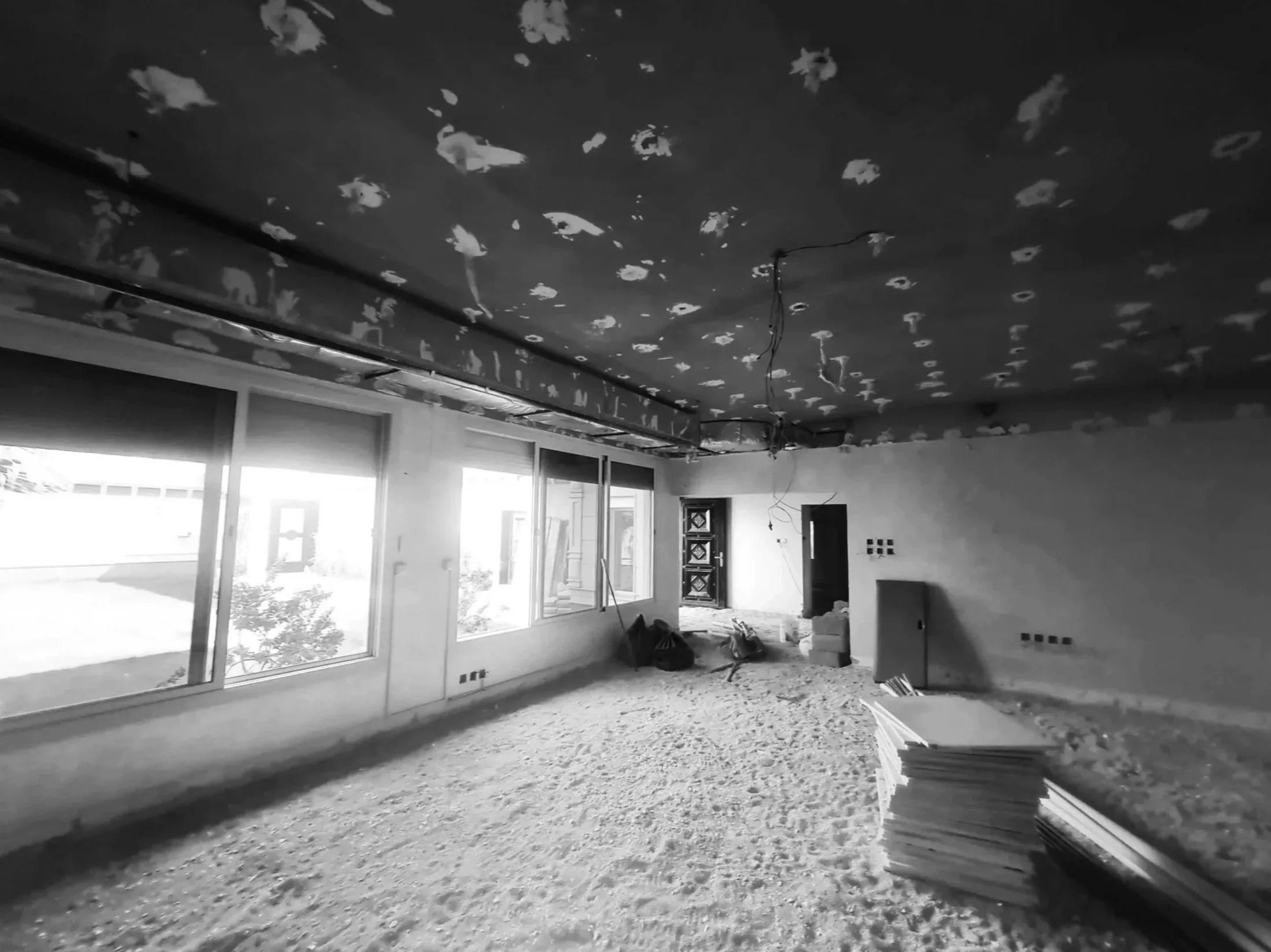 Interior room under construction with unfinished floors, ceiling, and large windows allowing natural light, construction materials and debris on the floor.