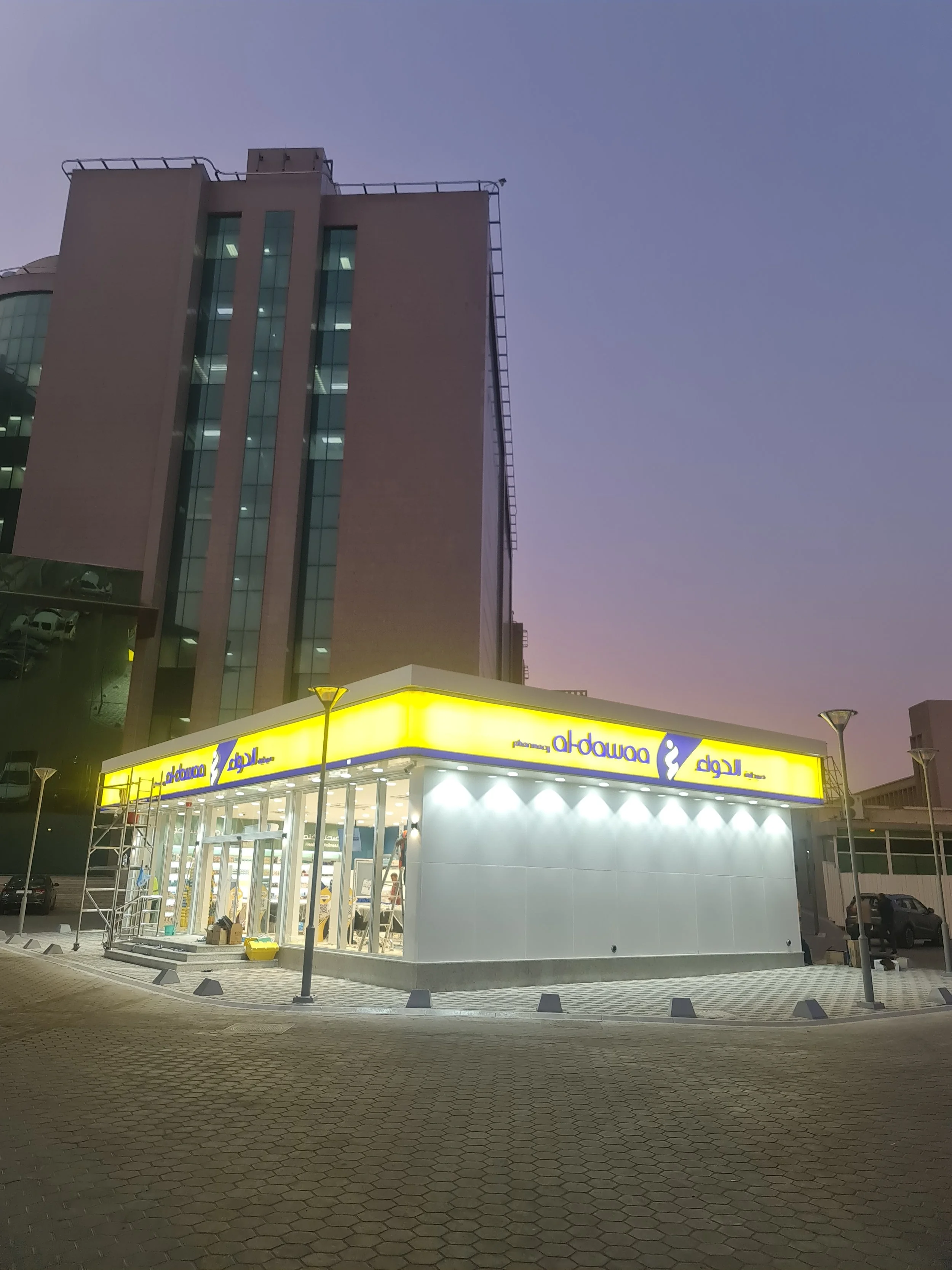 A brightly lit pharmacy store at dusk with a yellow sign displaying the pharmacy's name in English and Arabic, situated in an urban area with a tall building behind it and a paved parking lot.