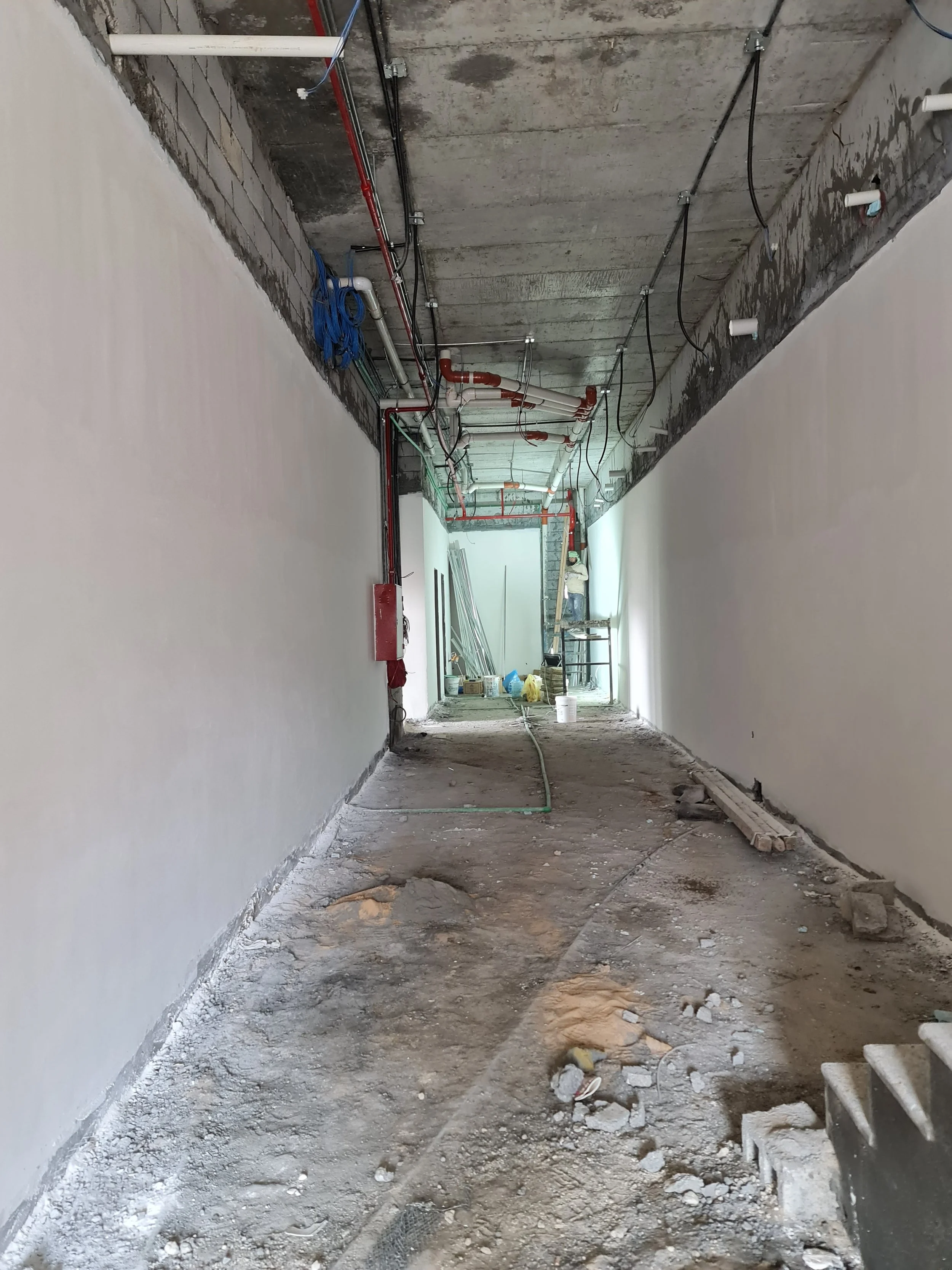Construction site hallway with exposed piping, electrical wiring, and unfinished floor.