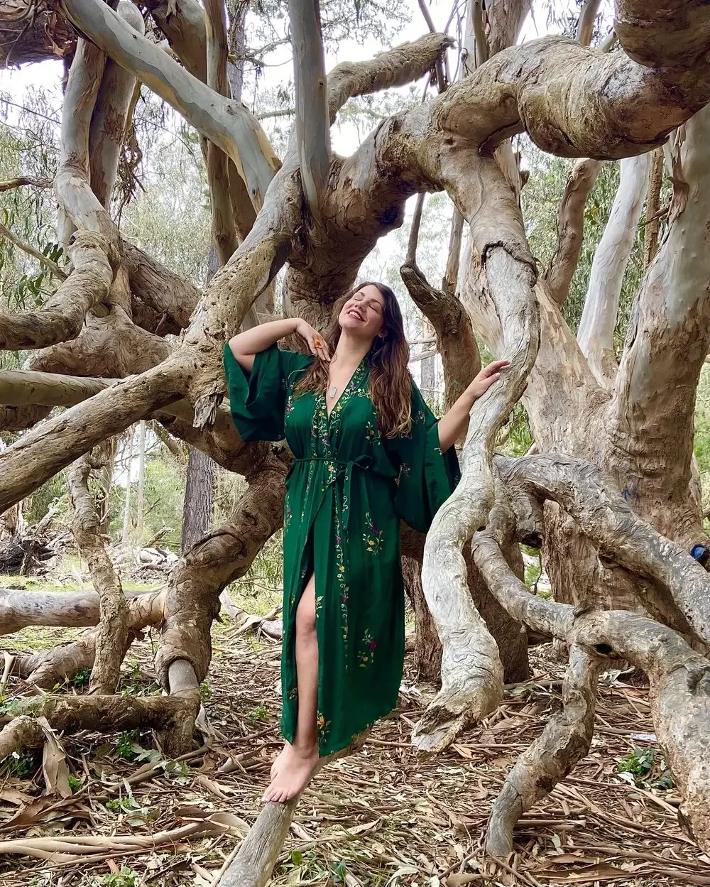 A woman in a long, green dress with floral embroidery is standing amidst the twisted branches of a large, ancient tree in a forest, smiling with her eyes closed and one hand touching her hair.