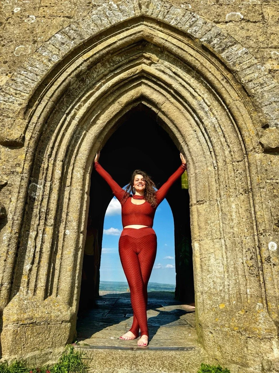 A woman in red textured athletic wear standing barefoot in a stone archway, smiling with arms raised, against a backdrop of blue sky and horizon.
