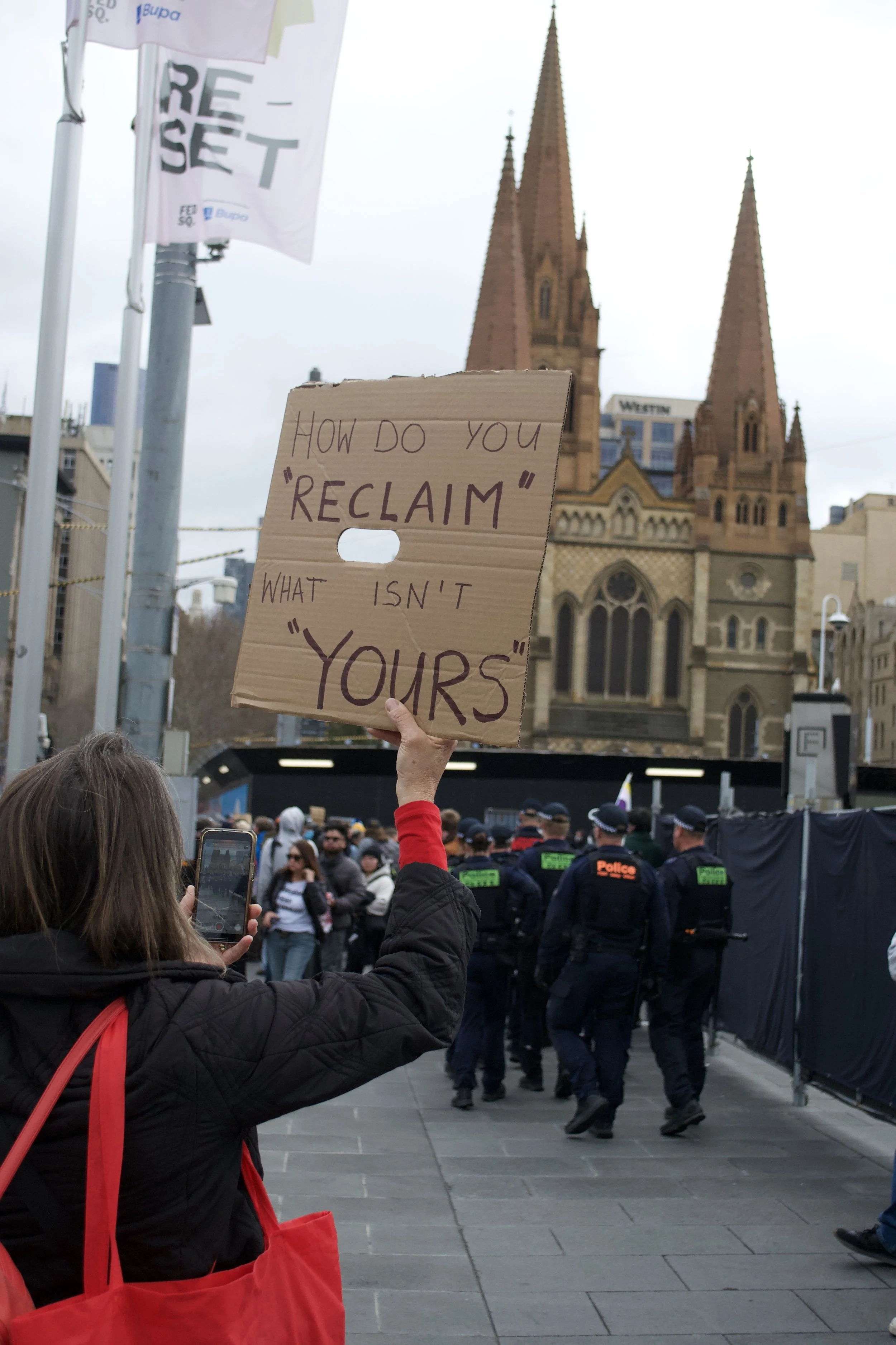 A counter protester holds up a sign directed towards Victoria Police 'How Do You "Reclaim" What Isn't "Yours"' at Fed Square.