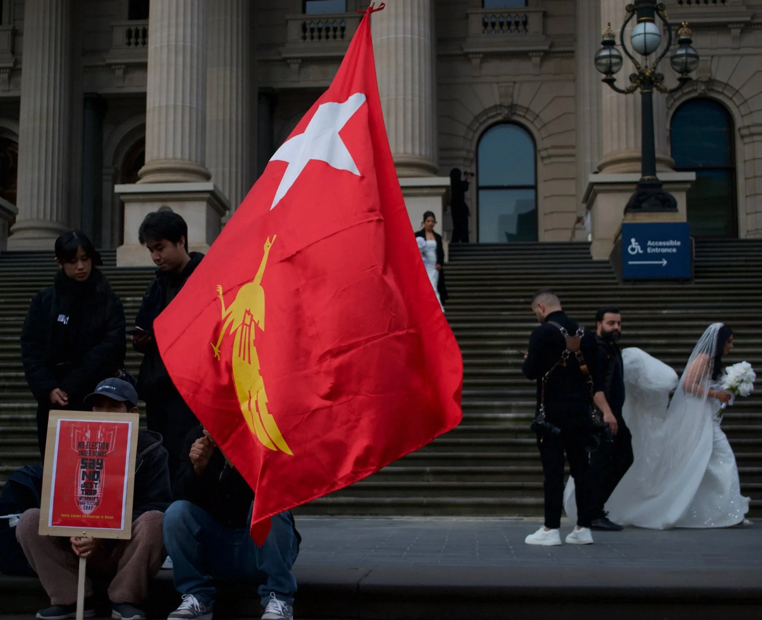 A protester waves the NLD flag next to another holding a sign 'No Election Under Bombs. Say No, Just Trap Myanmar's Sham Election Crap' whilst a wedding procession passes behind 06.09.2025
