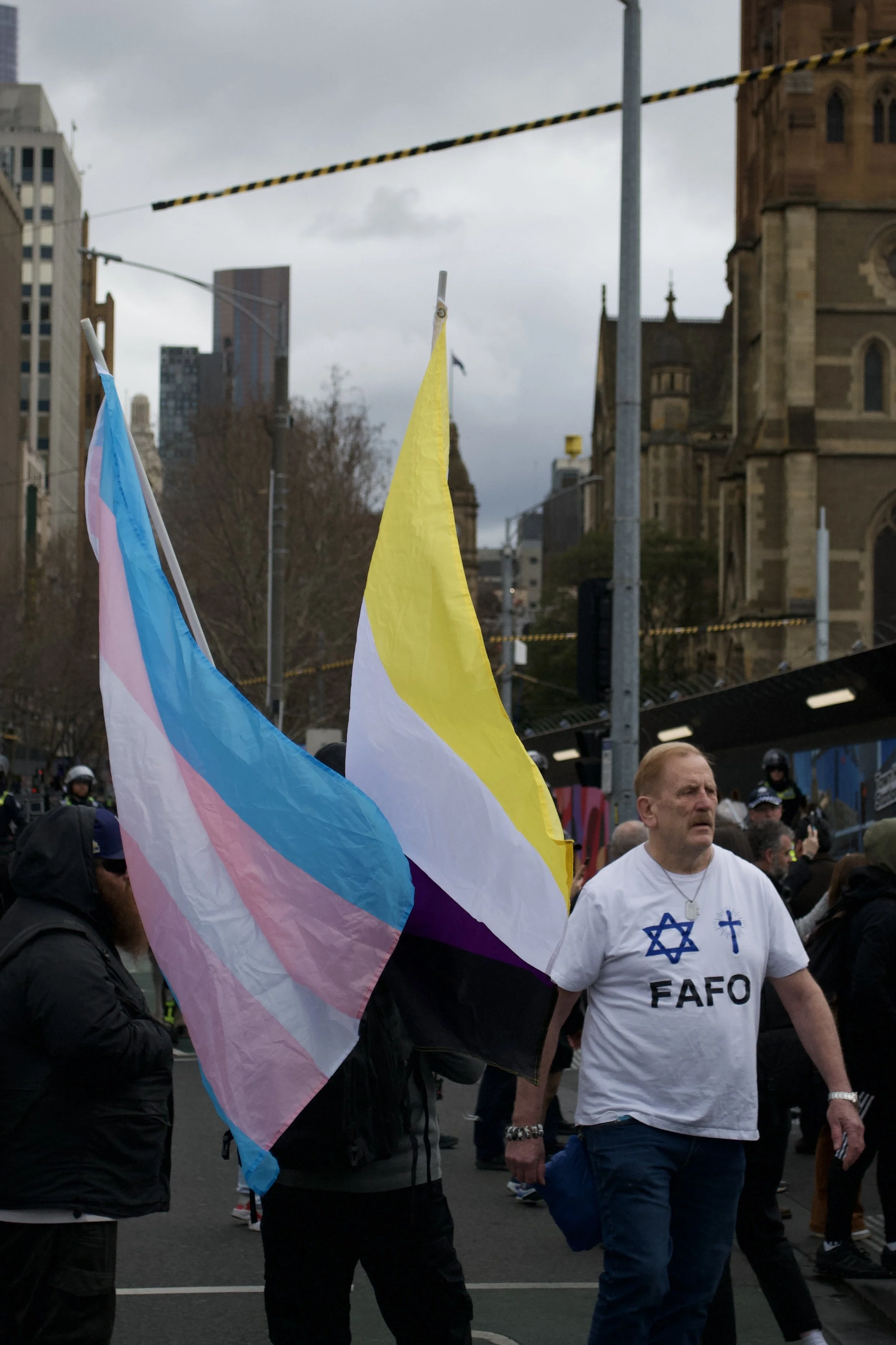 A man wearing a 'FAFO' t-shirt with the Star of David and a Crucifix on it walks past a counter protester holding the Transgender and Nonbinary flags at Fed Square.