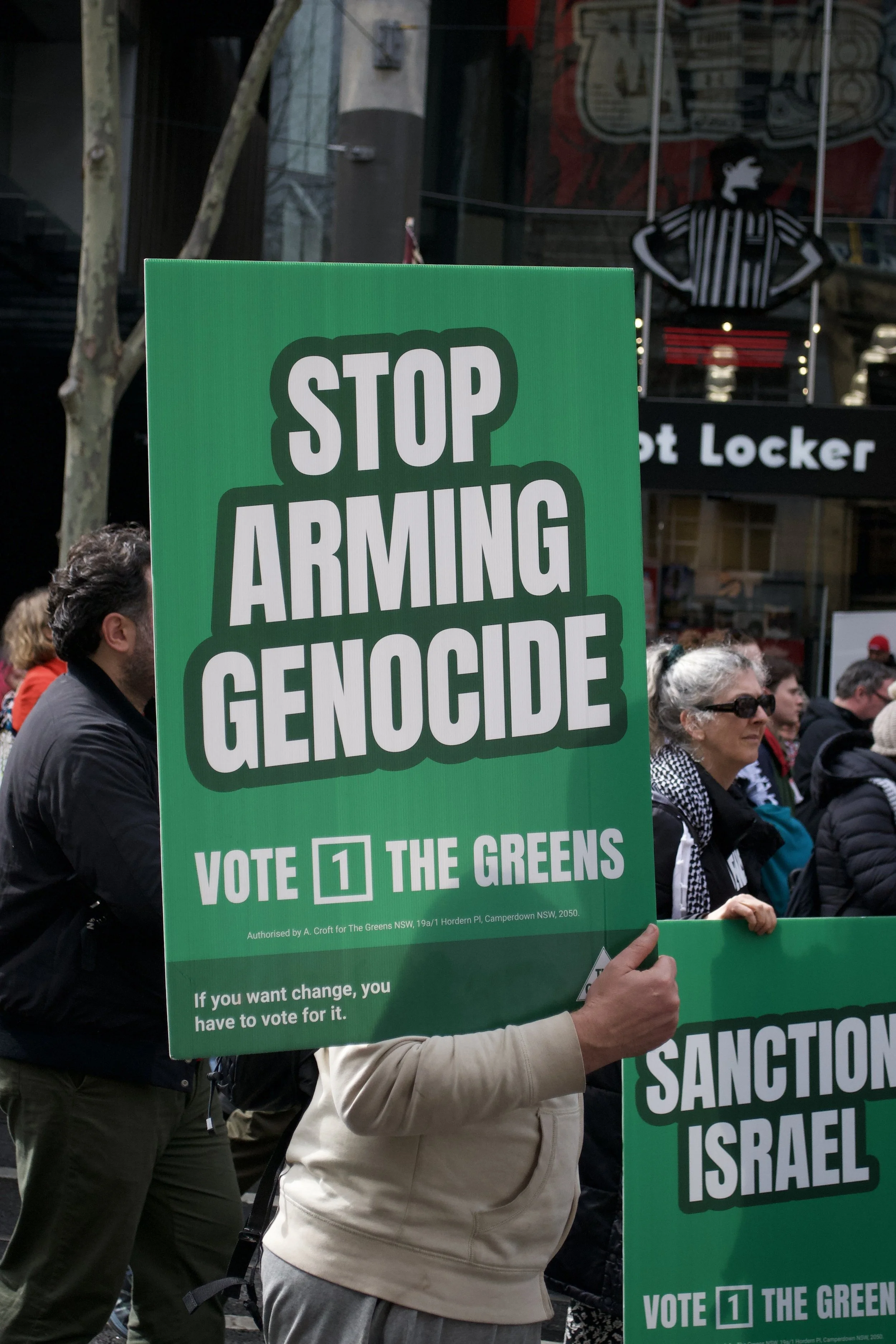 A counter protester holds up a Greens Party sign 'Stop Arming Genocide' out the front of QV on Swanston Street