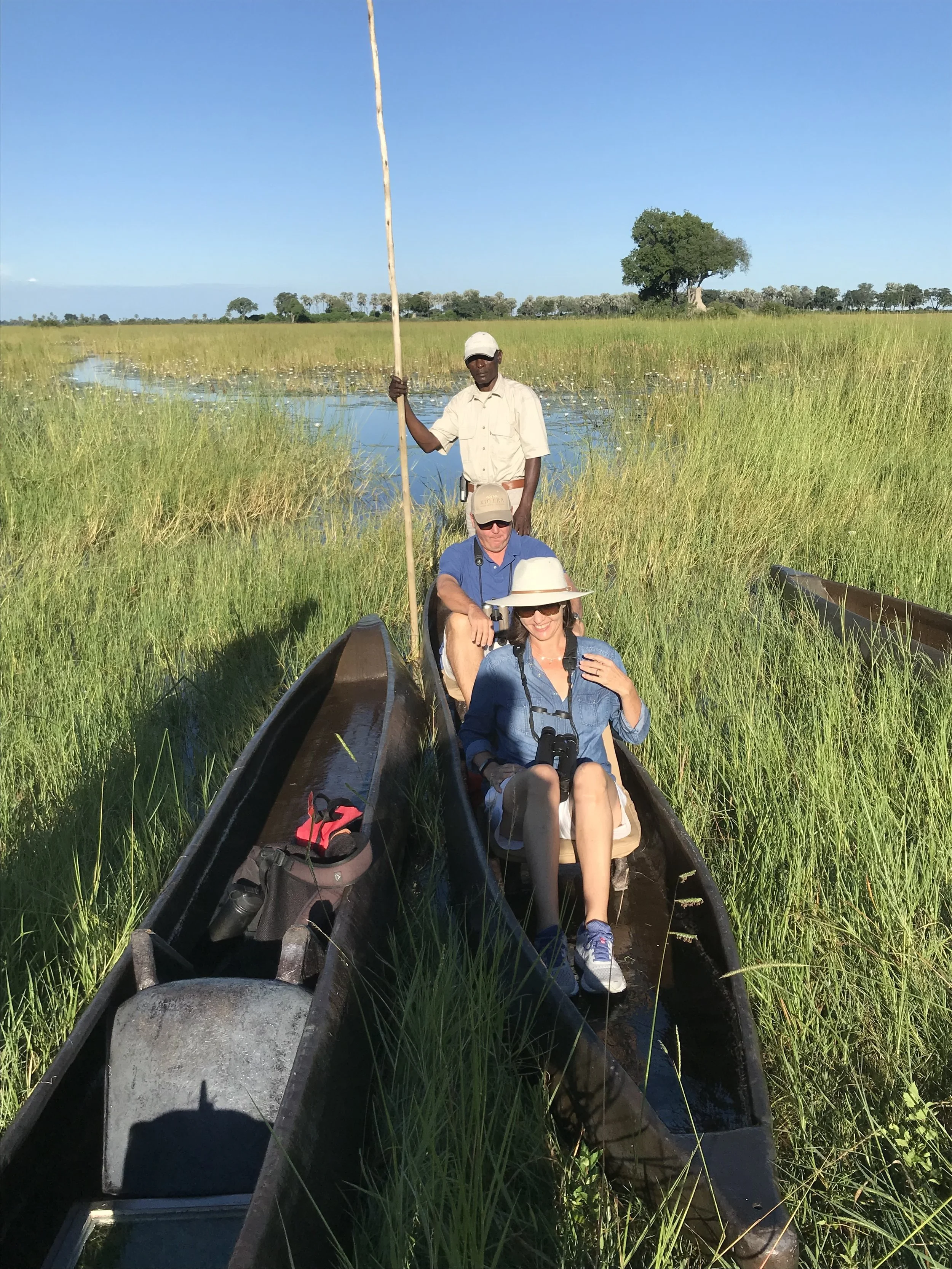 Three people on a boat in a marsh, two sitting and one standing, surrounded by tall grass and water, with clear blue sky and trees in the background.