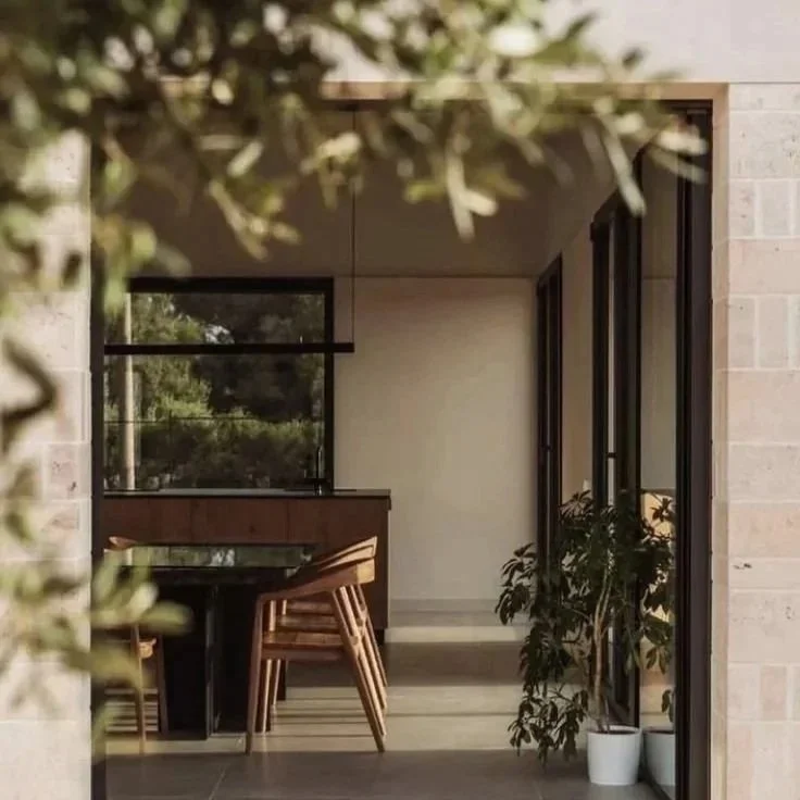 Modern dining area with wooden table, chairs, large glass doors, and potted plant, viewed through foliage.