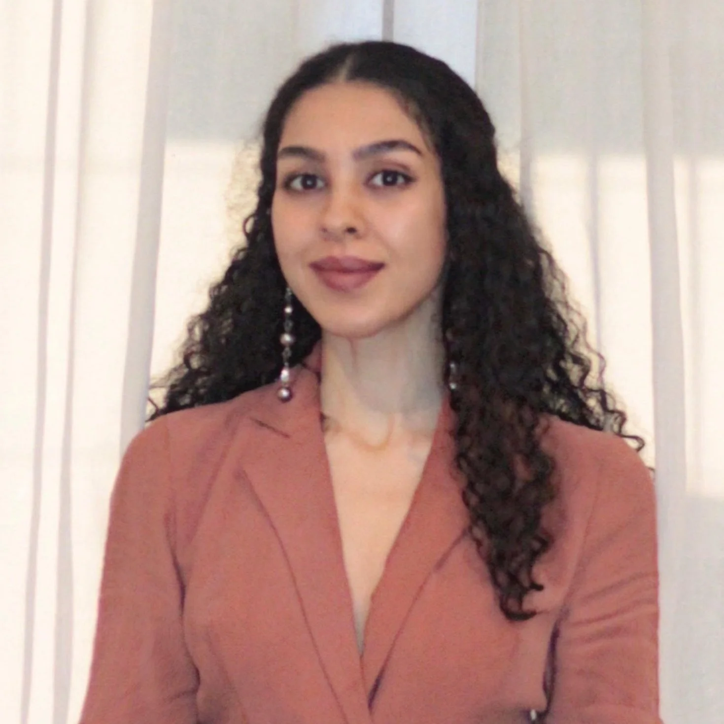 A woman with long curly dark hair wearing a rust-colored blazer and pearl earrings standing in front of a white wall with vertical panels.